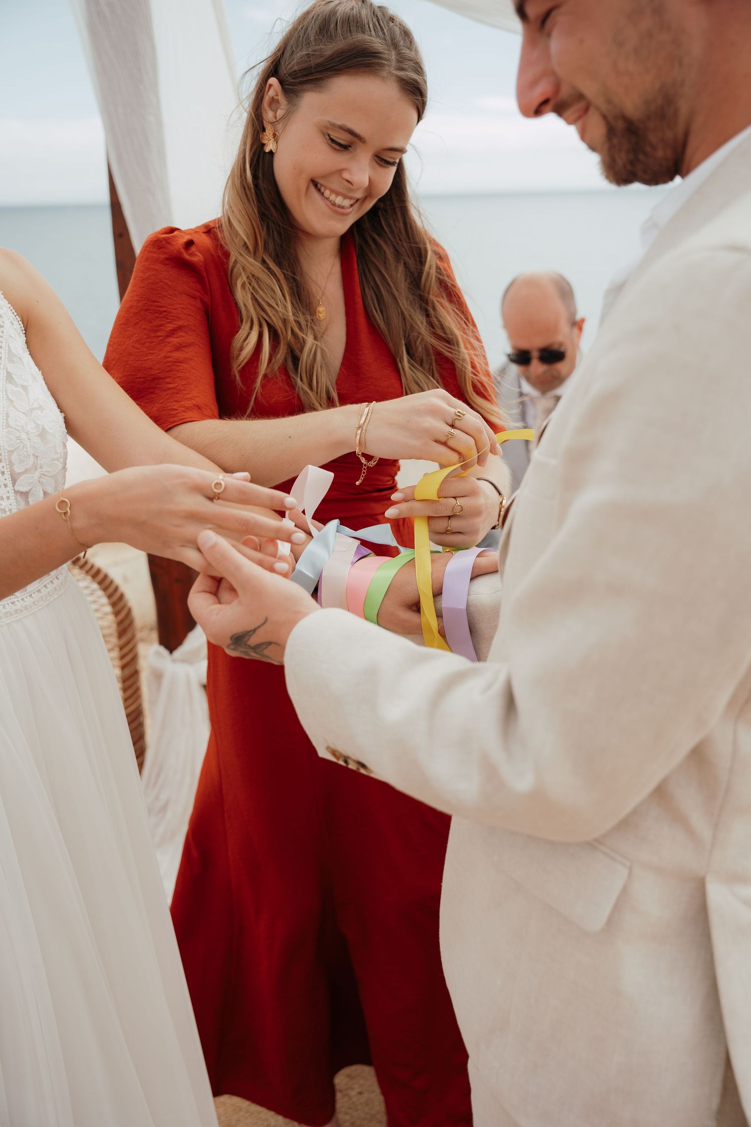 A woman in red dress smiles during a beach wedding handfasting ceremony with colorful ribbons.