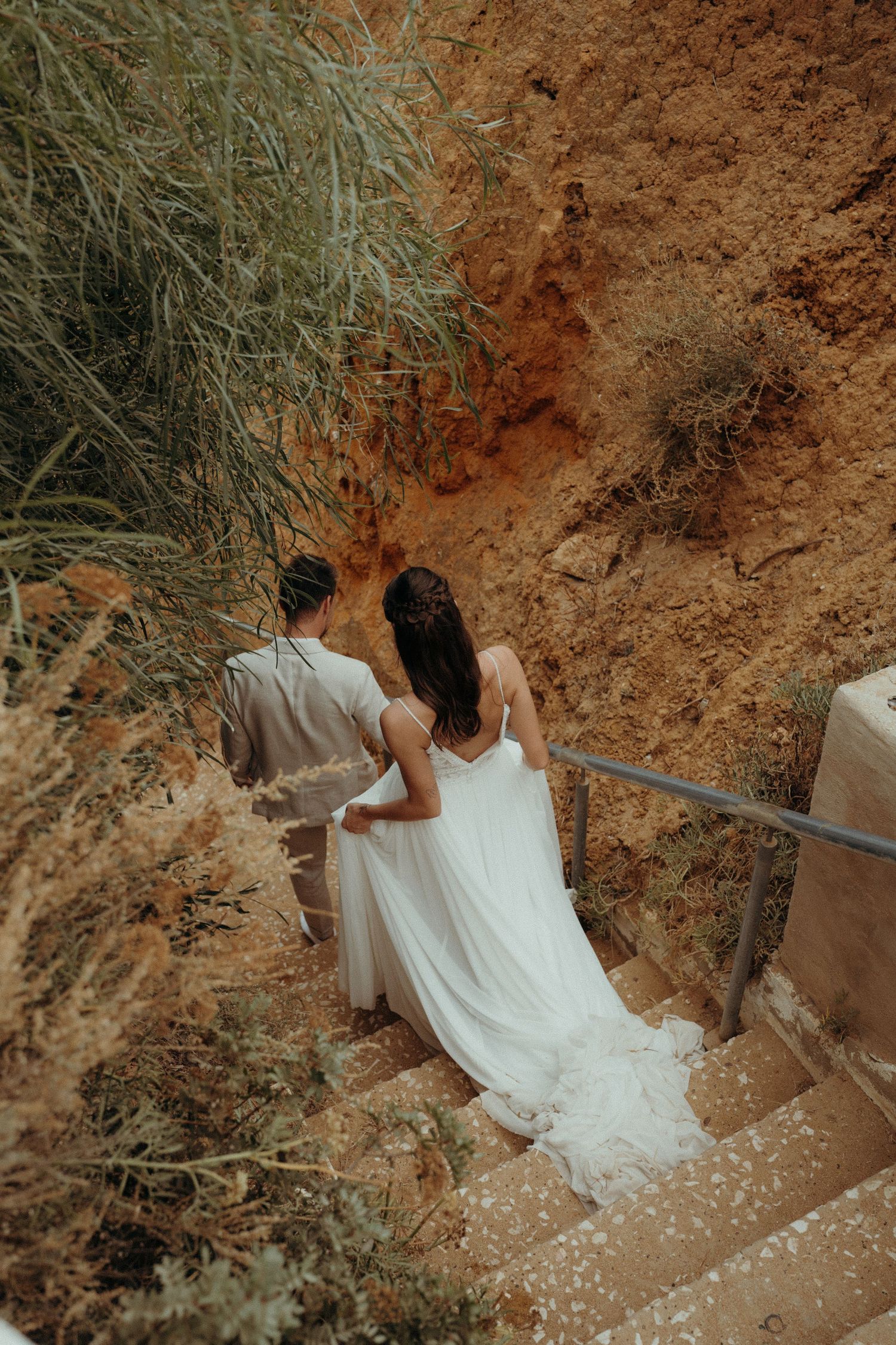 Bride and groom walking down stone steps surrounded by red cliffs and greenery on their wedding day.