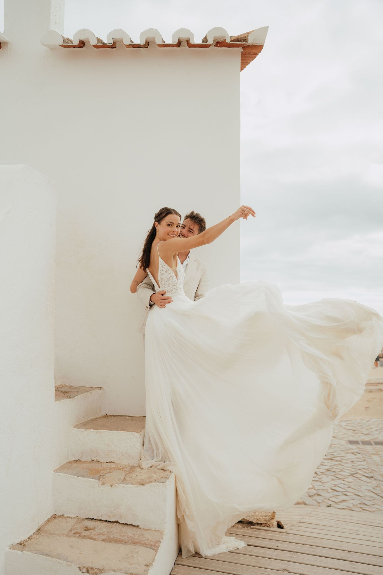 Bride in flowing white gown and groom embrace on sunlit Mediterranean steps, dress billowing in the breeze.