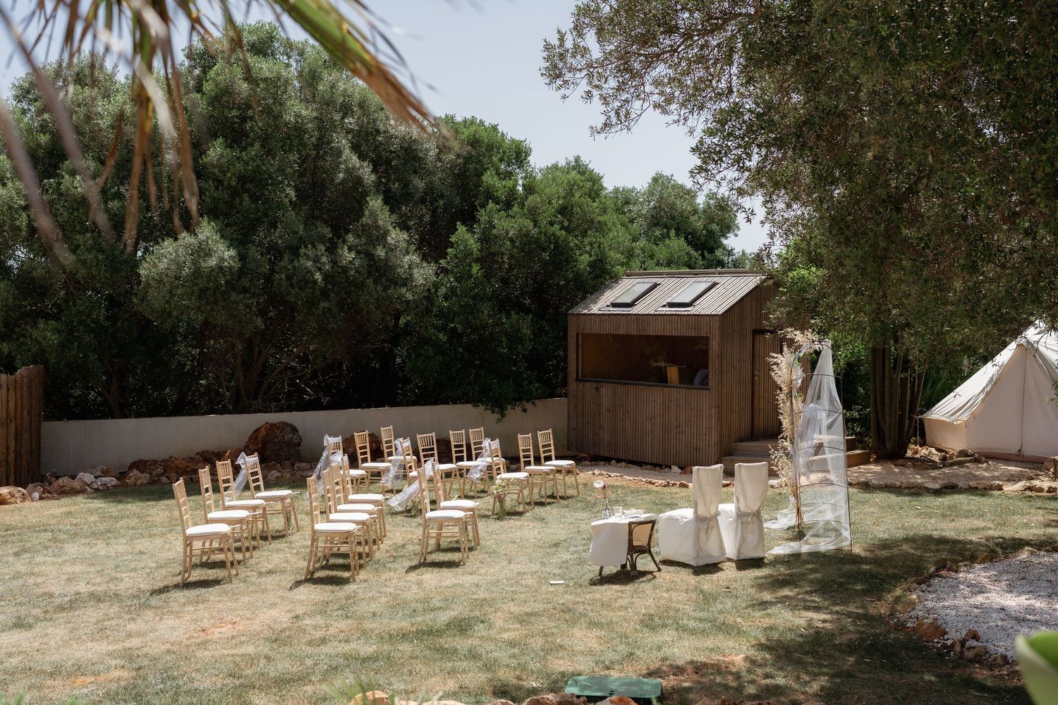 Outdoor wedding ceremony setup with wooden chairs on grass, a rustic wooden structure, and a white tent among trees.