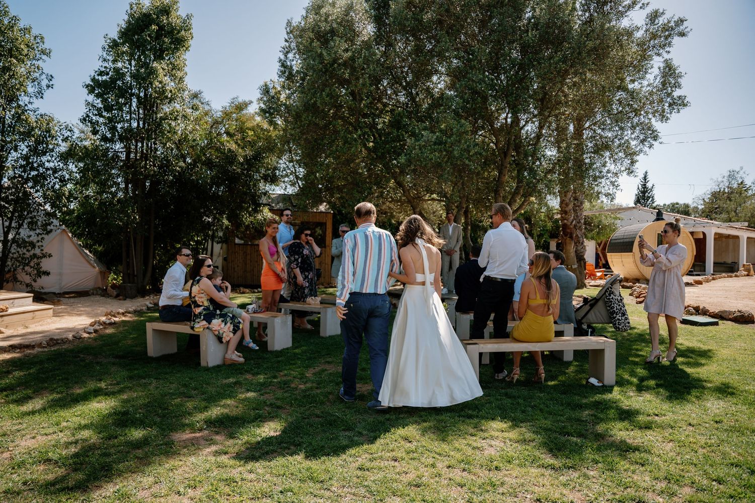 Bride and groom stand with wedding guests on green lawn beneath tall trees at outdoor ceremony.