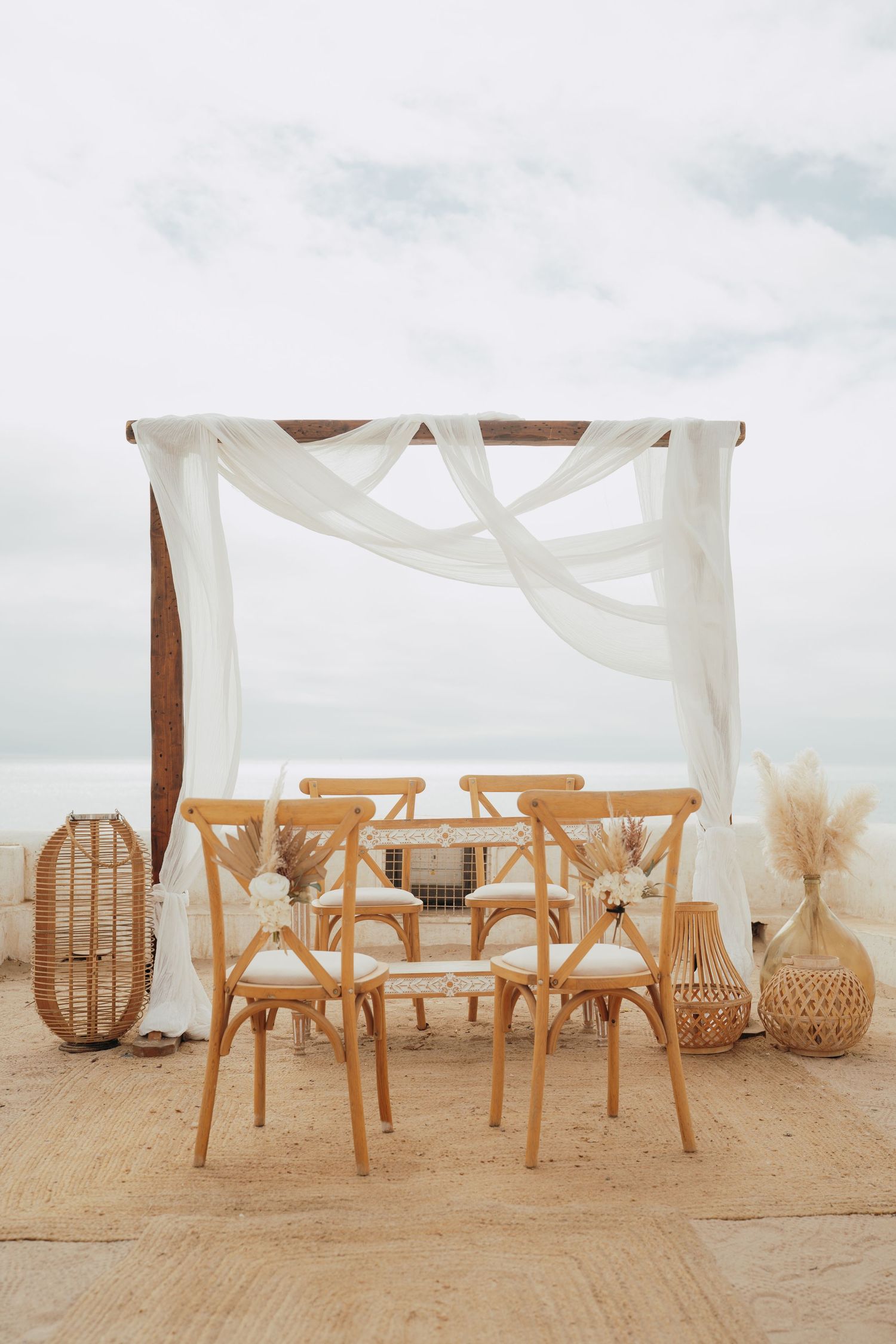 Boho beach wedding setup with wooden chairs, canopy draped in white fabric, rattan decor, and pampas grass by the ocean.