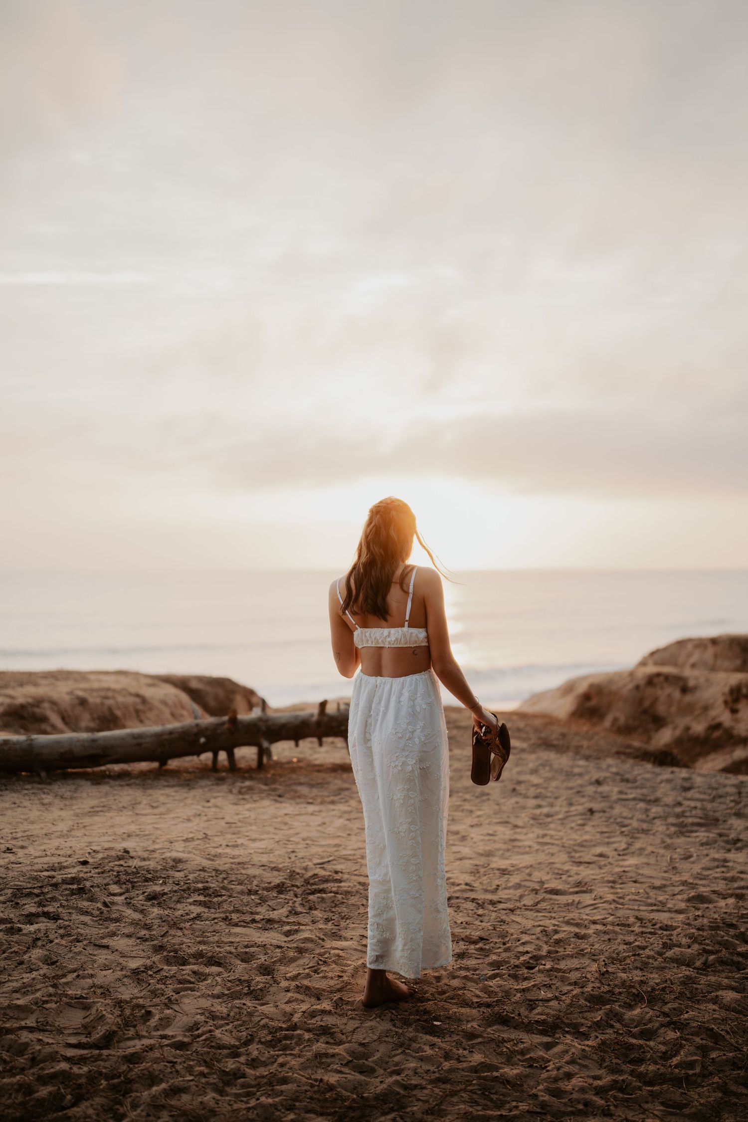 Woman in white dress holding shoes on sandy beach at sunset, facing the ocean with golden light.