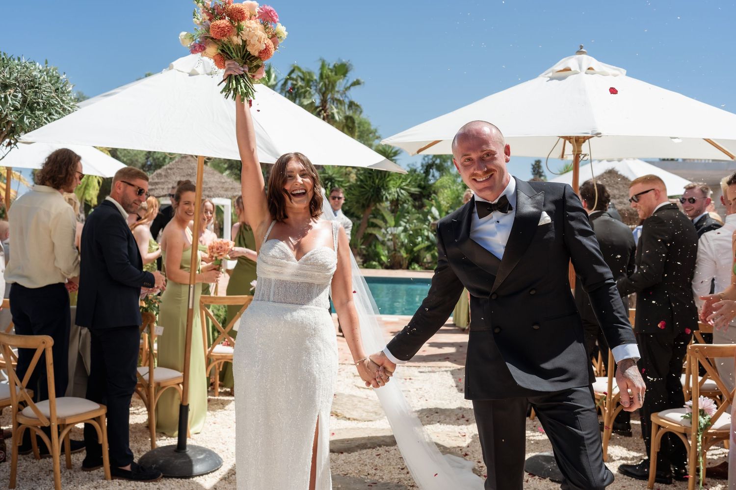 Joyful bride and groom walk hand-in-hand after outdoor wedding ceremony, bride raising bouquet in celebration.