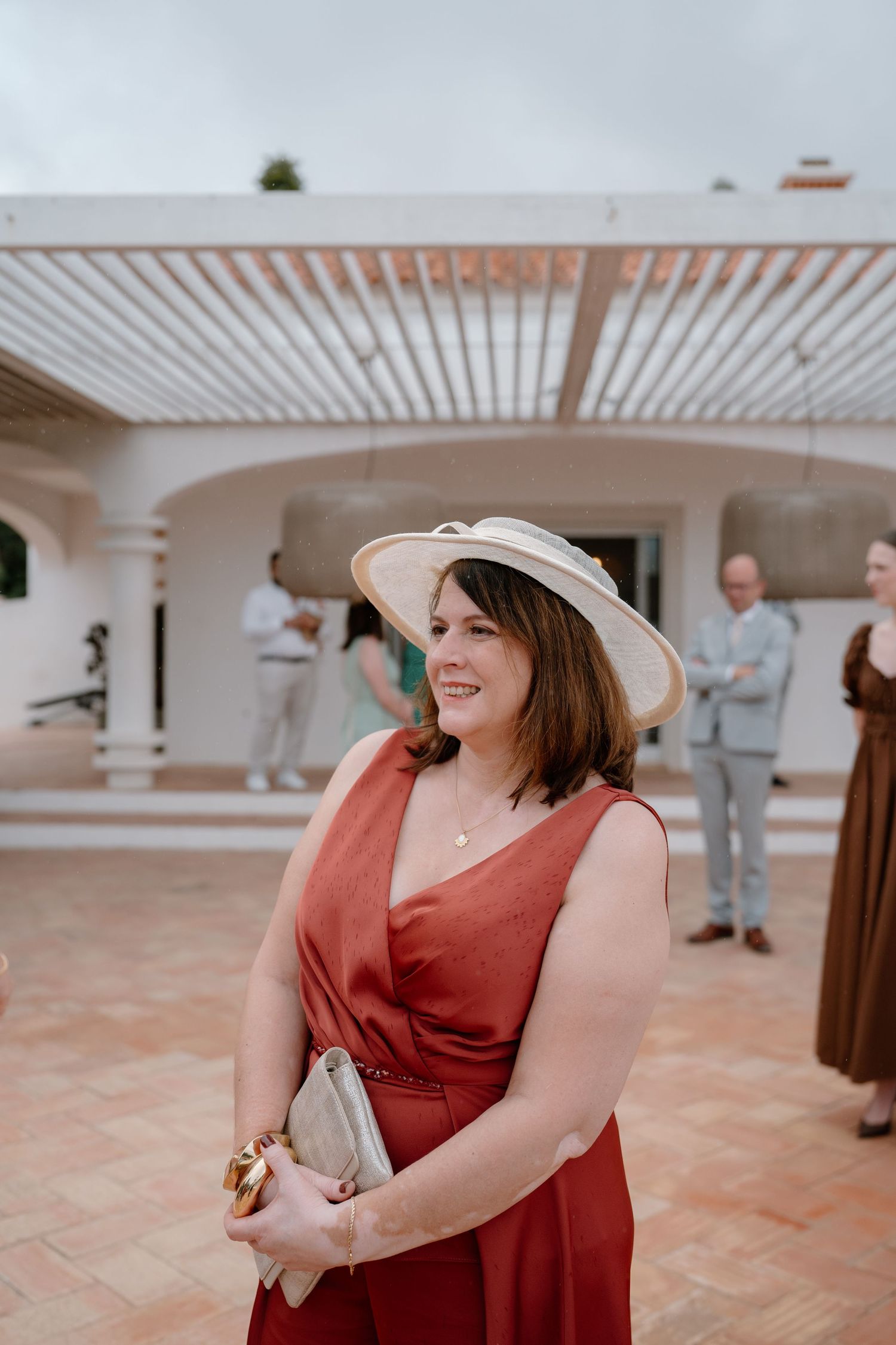 Woman in red dress and wide-brim hat smiling at outdoor wedding reception on terrace.