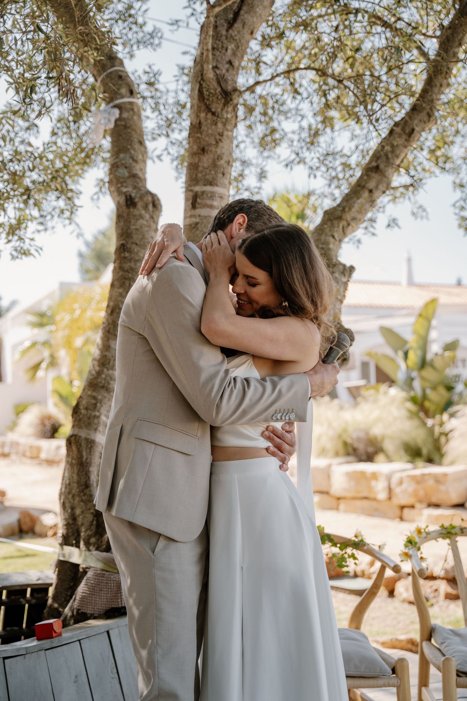 Bride and groom share a heartfelt embrace under olive trees during their outdoor wedding ceremony.