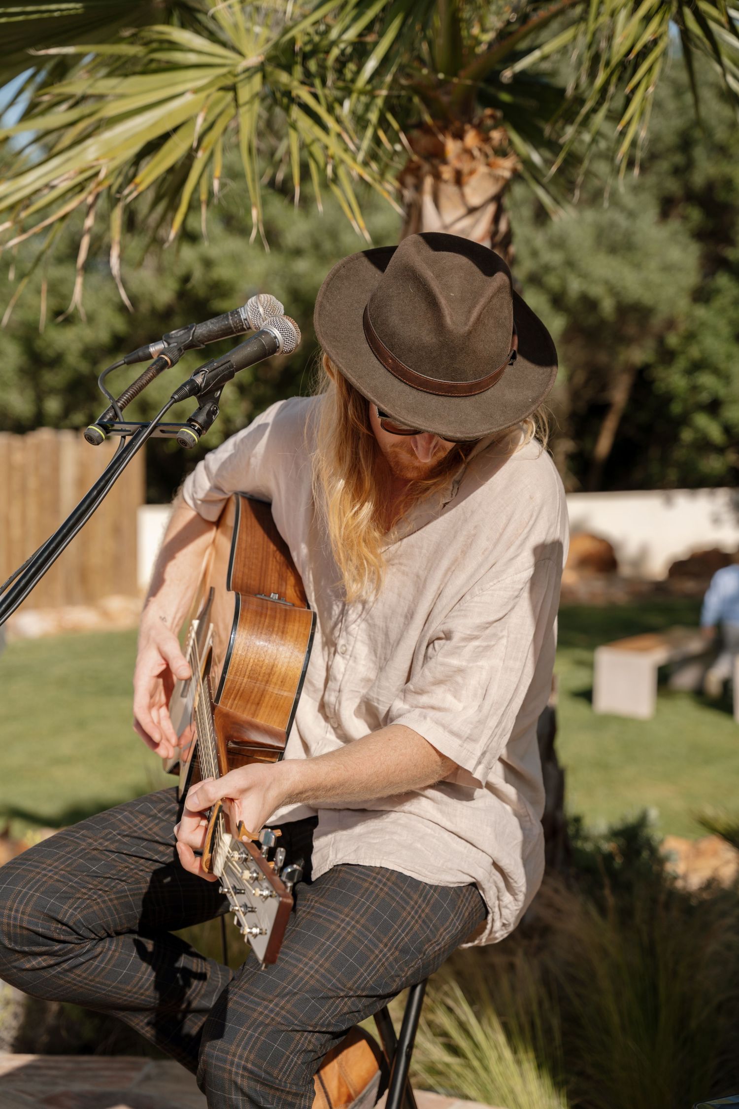 Musician with a cowboy hat plays acoustic guitar outdoors at a garden wedding.