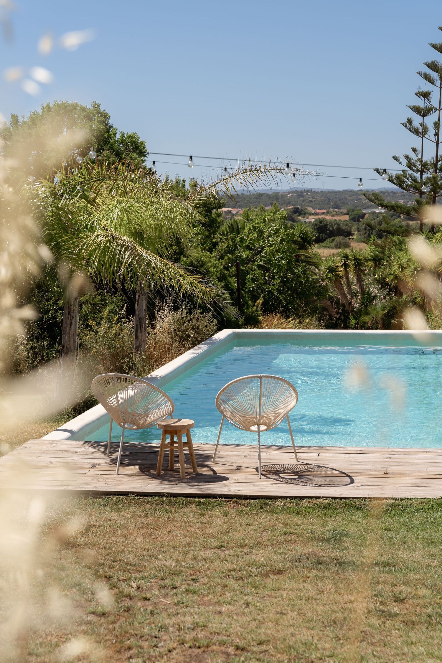 Two wicker chairs on a wooden deck beside a serene outdoor pool surrounded by lush greenery under a clear blue sky.