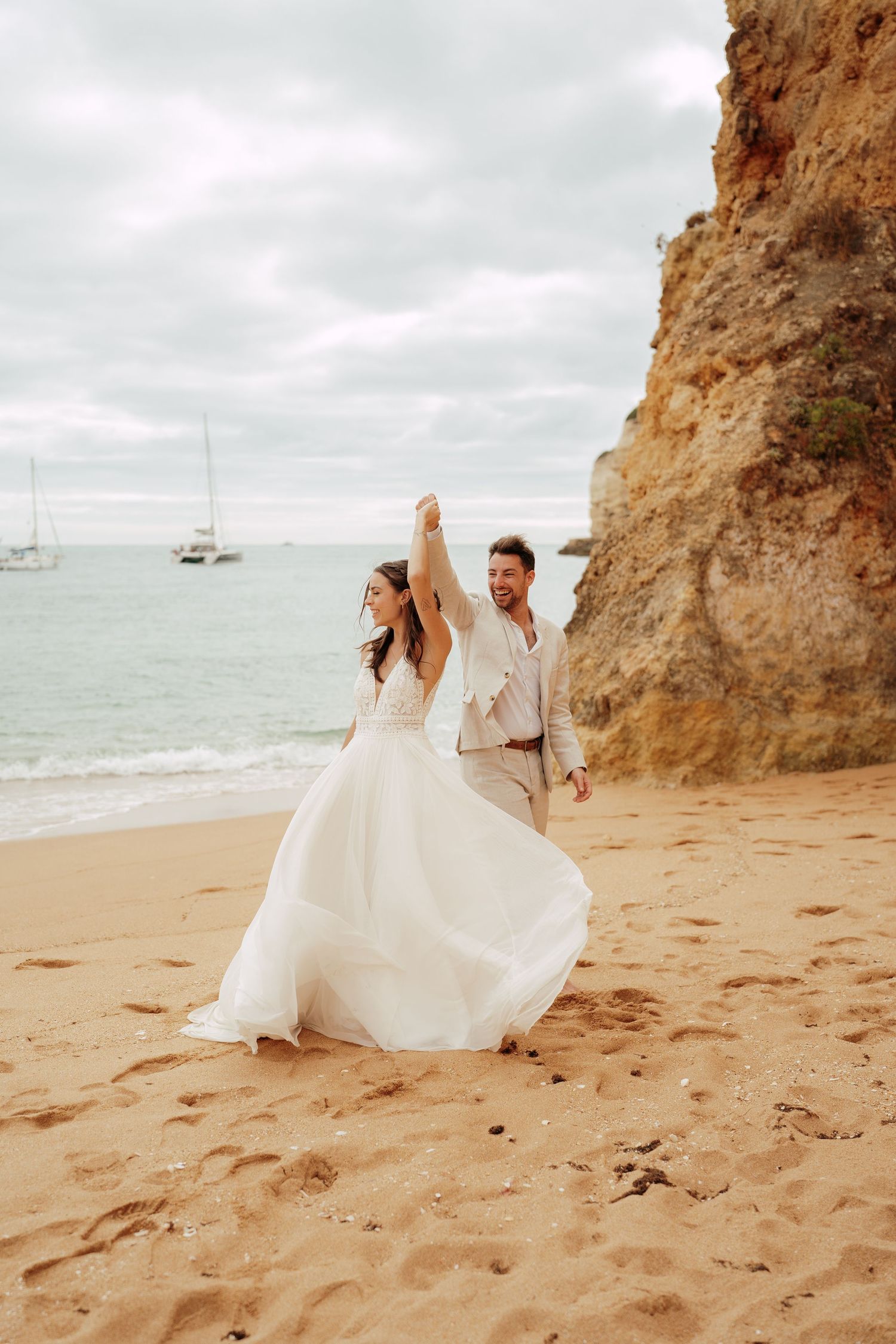 Bride and groom holding hands on sandy beach with rocky cliffs and ocean backdrop on their wedding day.