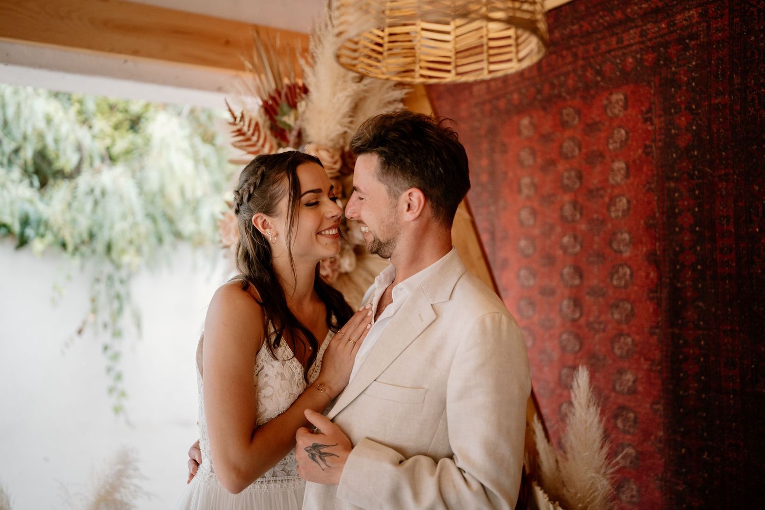 A couple in wedding attire share an intimate moment, smiling closely together against a boho-styled backdrop.
