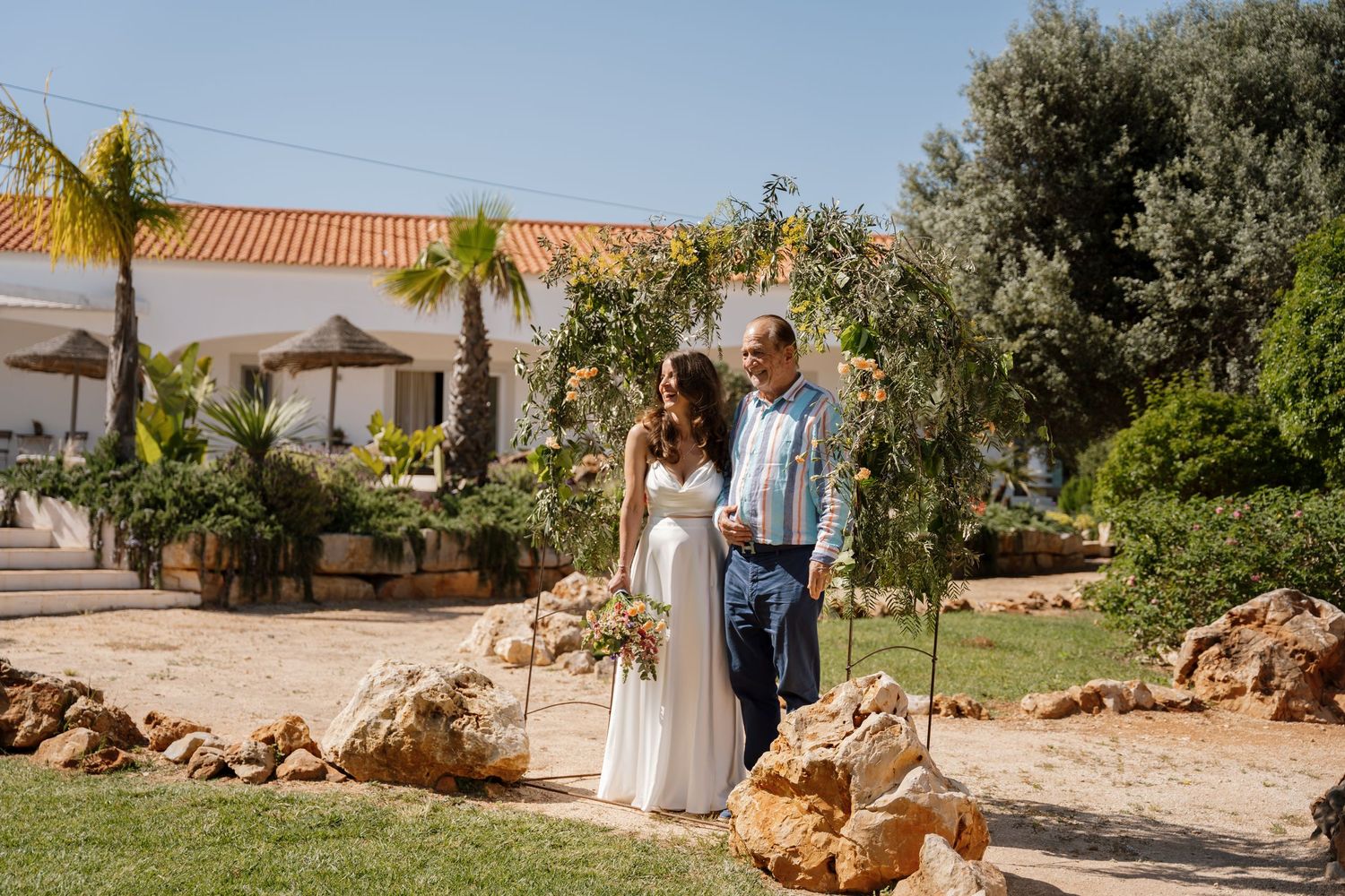 Wedding couple stands near a floral arch in a rocky garden with a white Mediterranean villa backdrop.