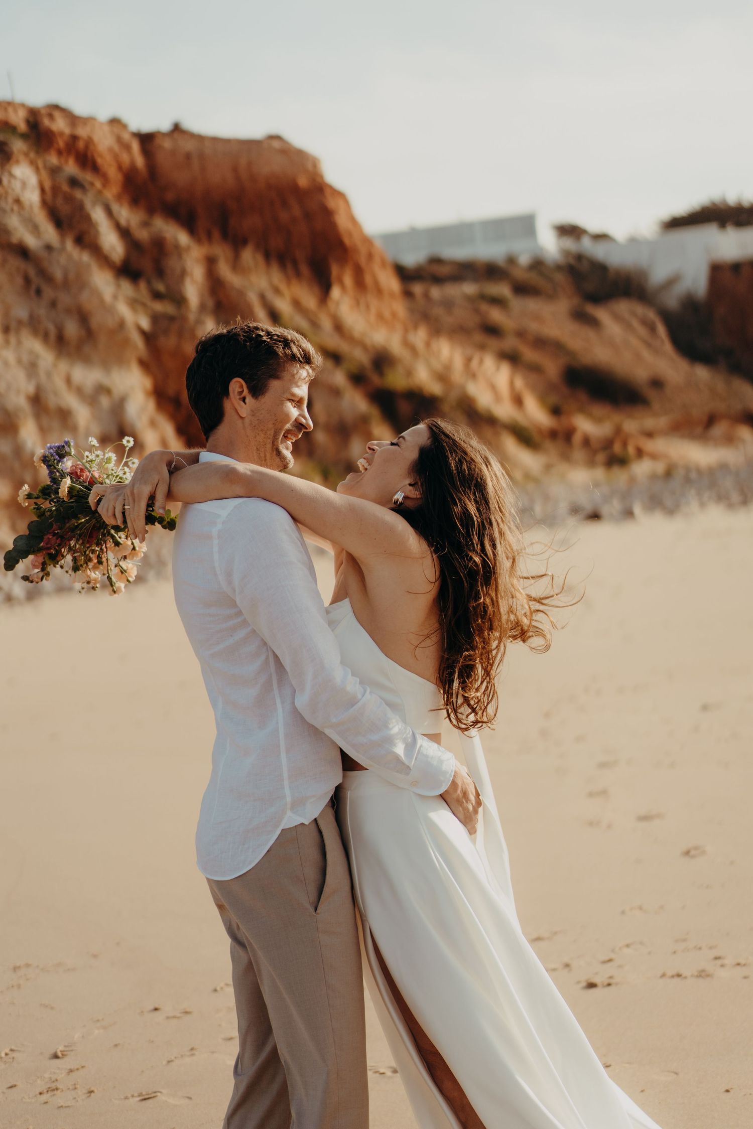 Laughing bride and groom embrace joyfully on a sandy beach with golden cliffs glowing in the background.