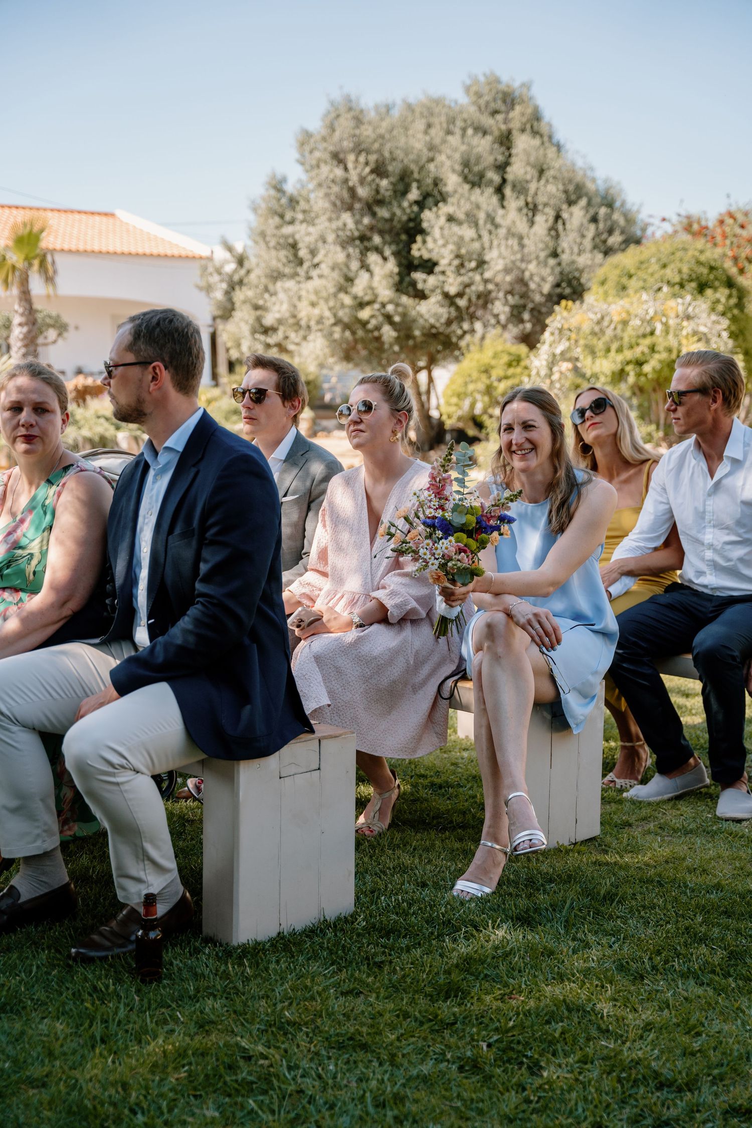 Smiling wedding guests in summer attire sit on wooden benches during outdoor garden wedding ceremony.