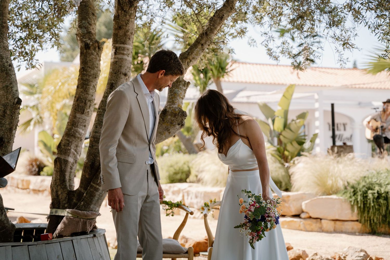 Bride and groom look down together at colorful bouquet near olive trees at sunny outdoor wedding.