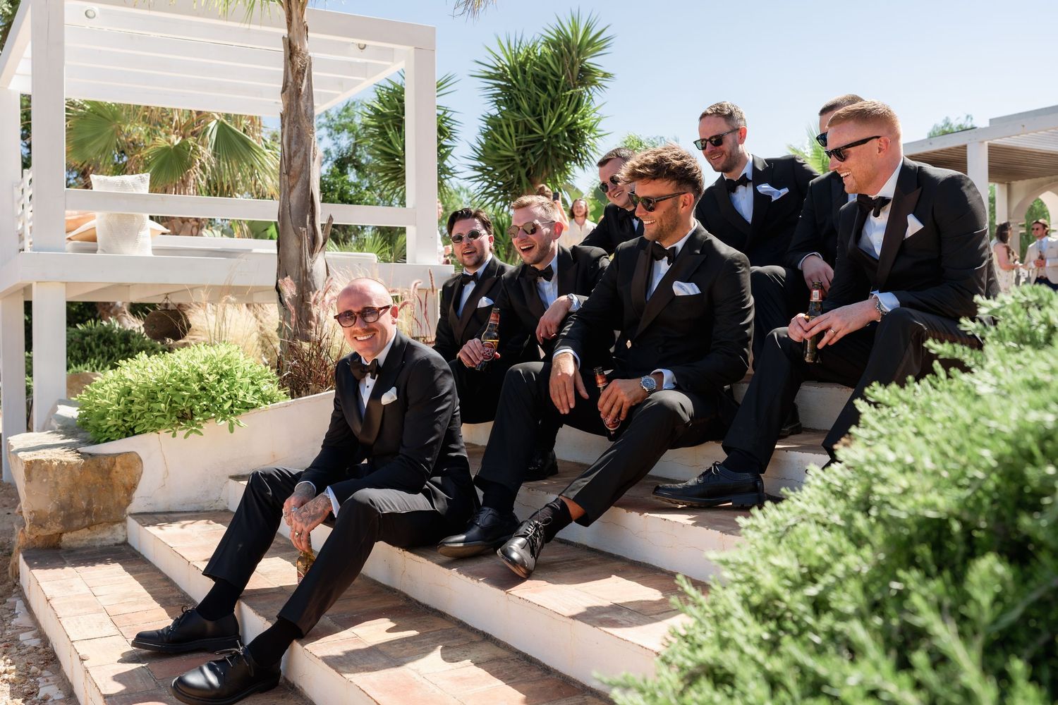Groomsmen in black tuxedos and bow ties relaxing on outdoor steps at a sunny wedding venue.