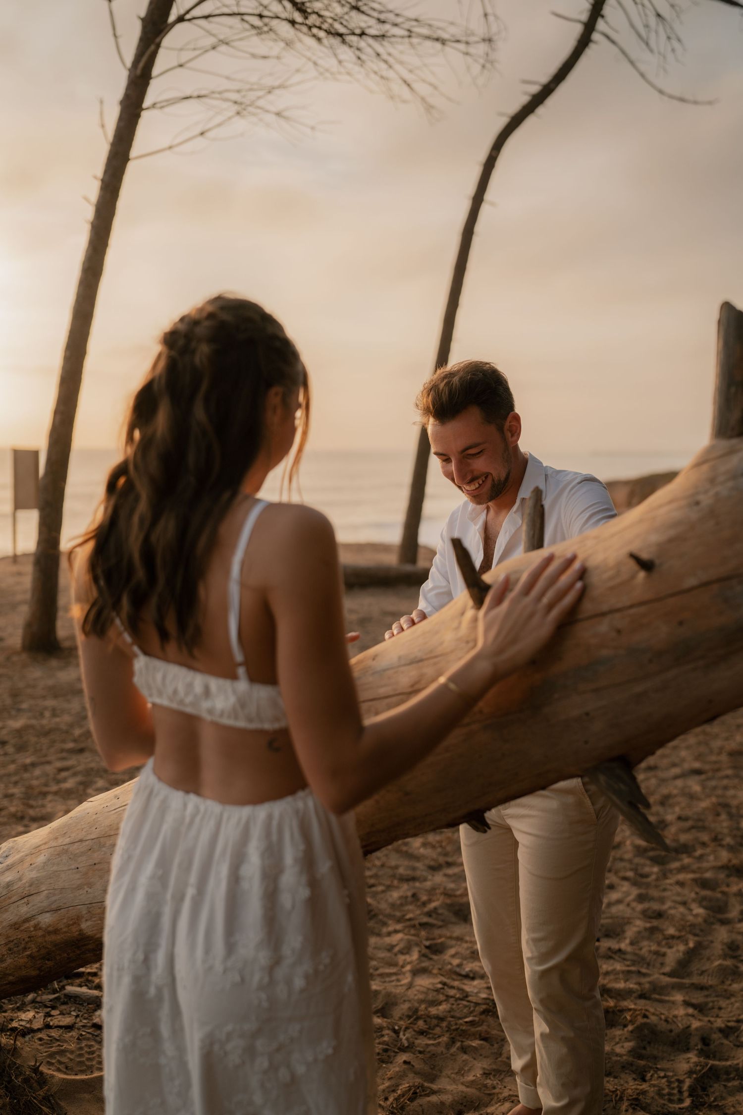 Couple in white attire sharing a romantic moment by a driftwood log on a sandy beach at golden sunset.