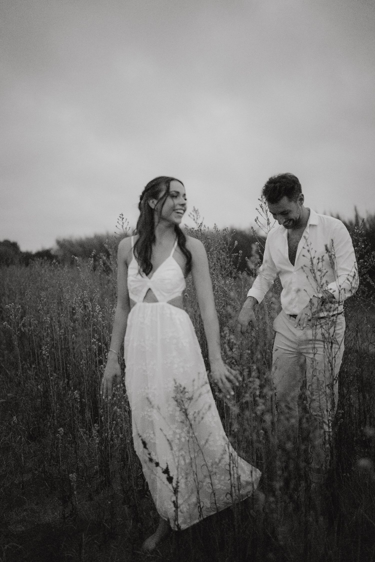 Black and white photo of a smiling bride in white dress holding hands with groom walking through wildflower field.