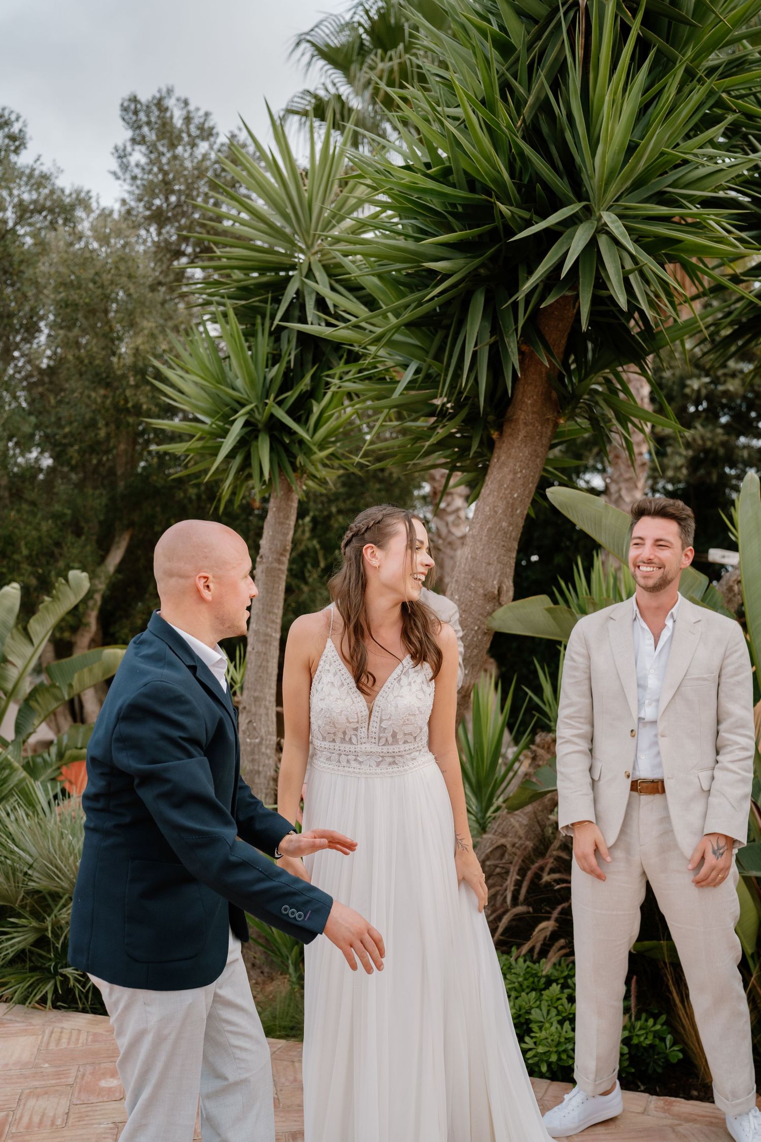 Bride in white lace gown smiles with two suited grooms outdoors near tropical palm trees at a wedding ceremony.