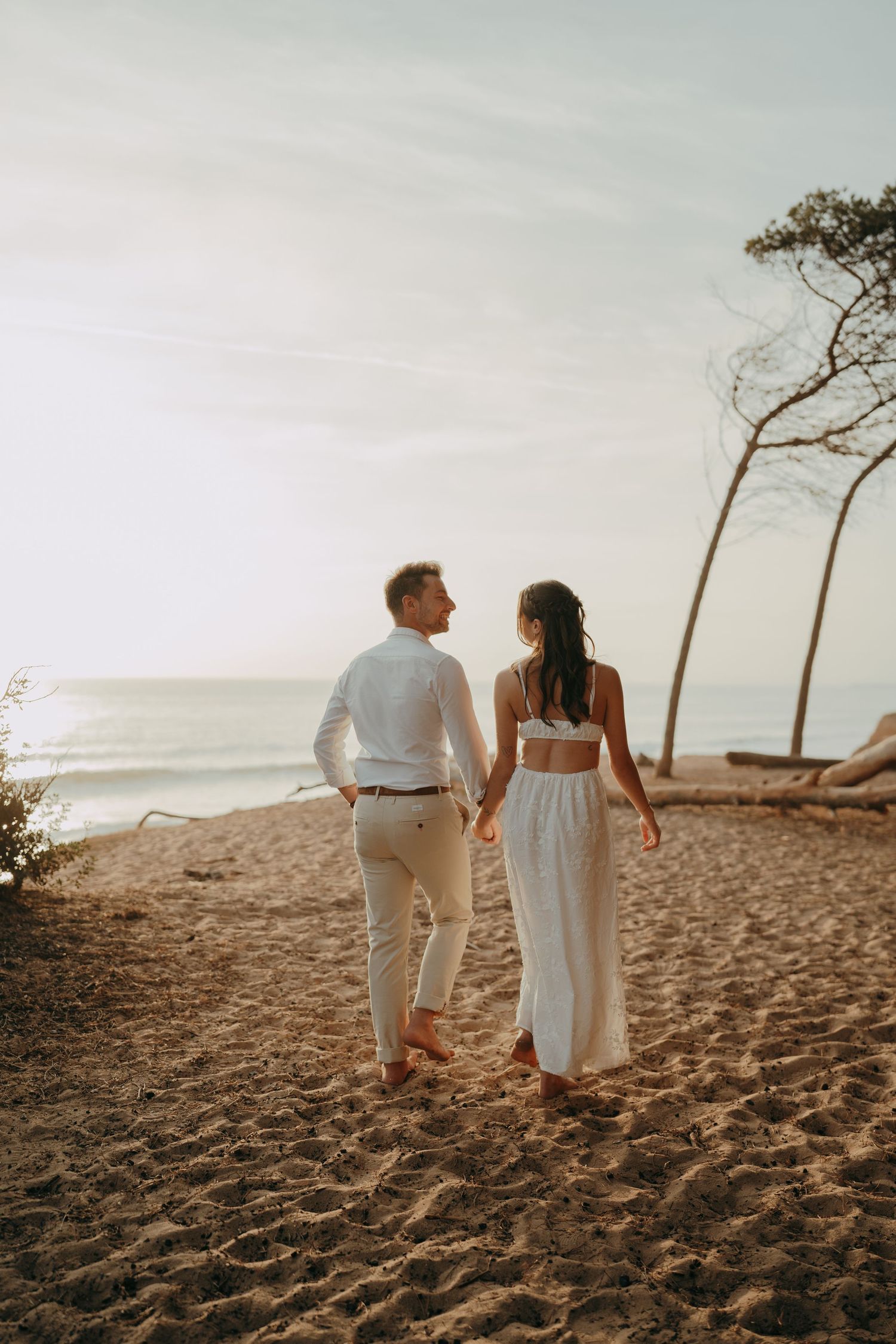 Couple in white outfits holding hands while walking on a sandy beach at sunset near the ocean.