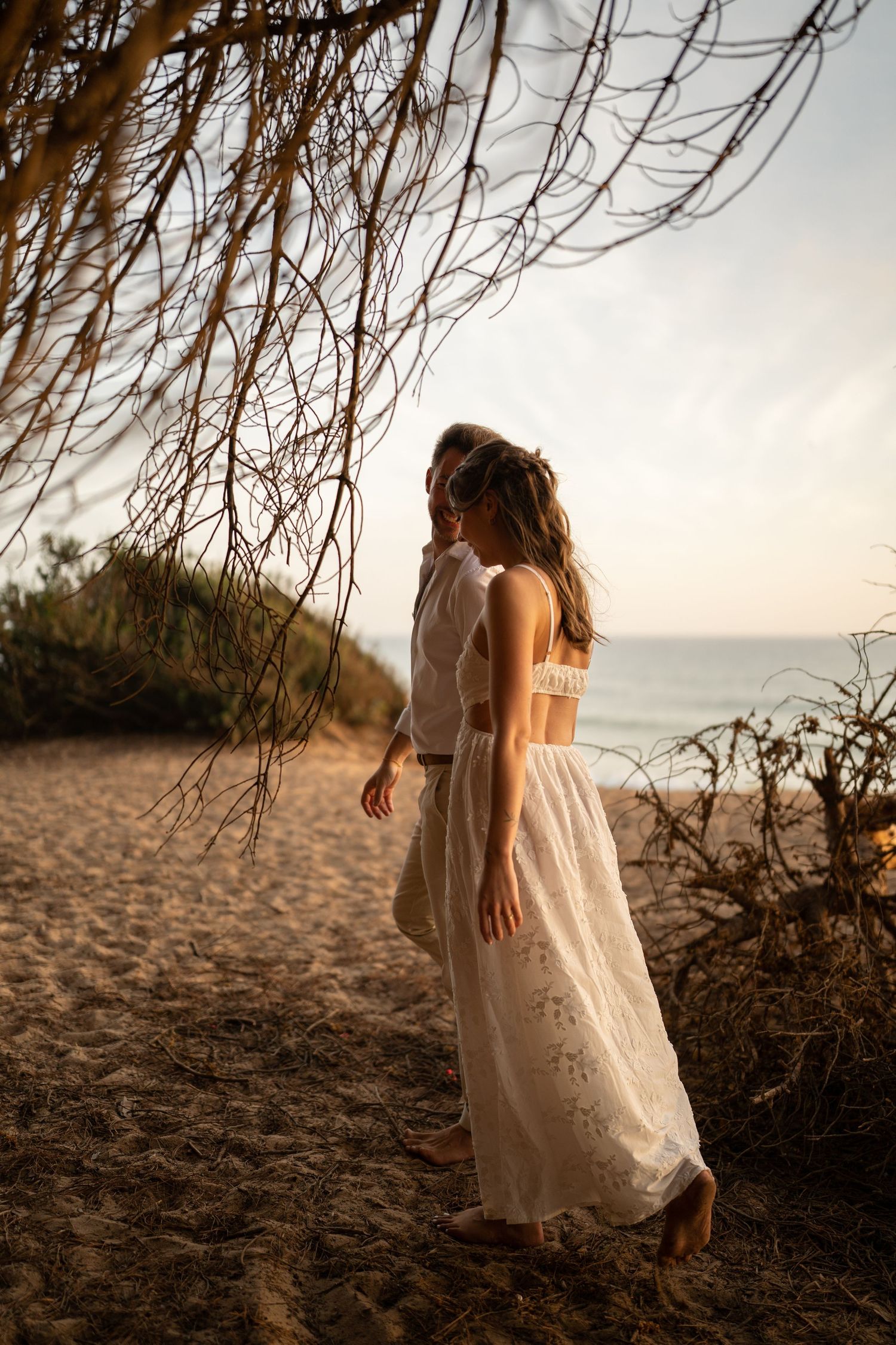 A woman in a white lace maxi dress stands on a sandy beach path at golden hour, framed by bare branches.