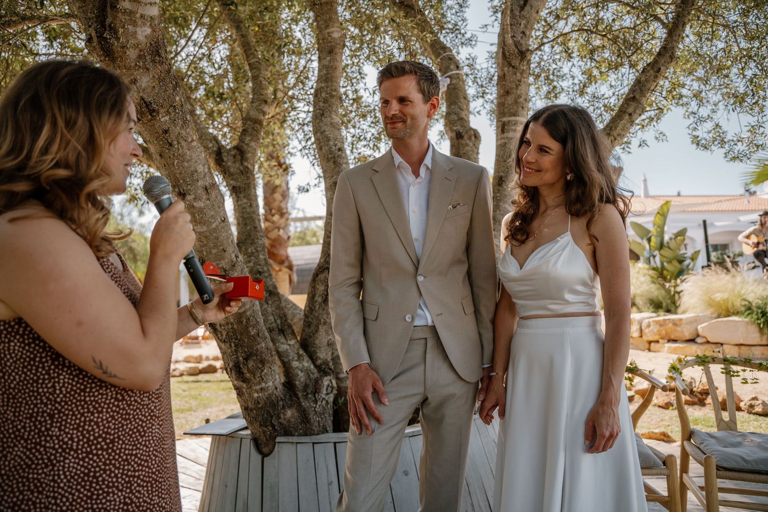 Officiant speaks to smiling couple during outdoor wedding ceremony under olive trees.