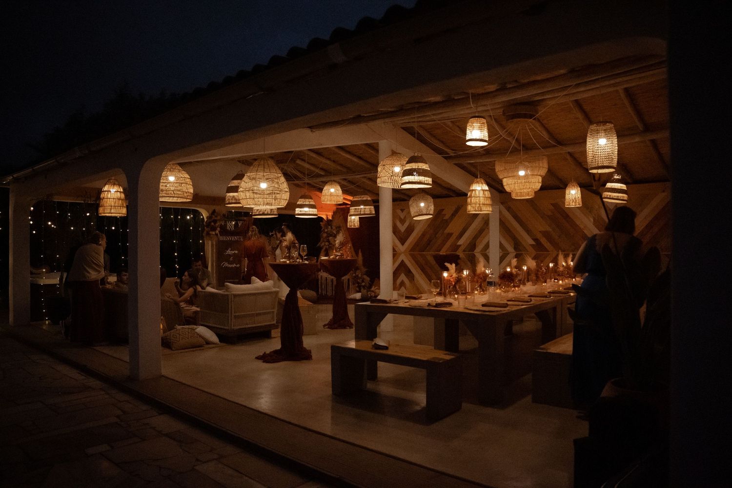 Warmly lit outdoor dining pavilion at night with wicker pendant lights and long tables set for evening guests.