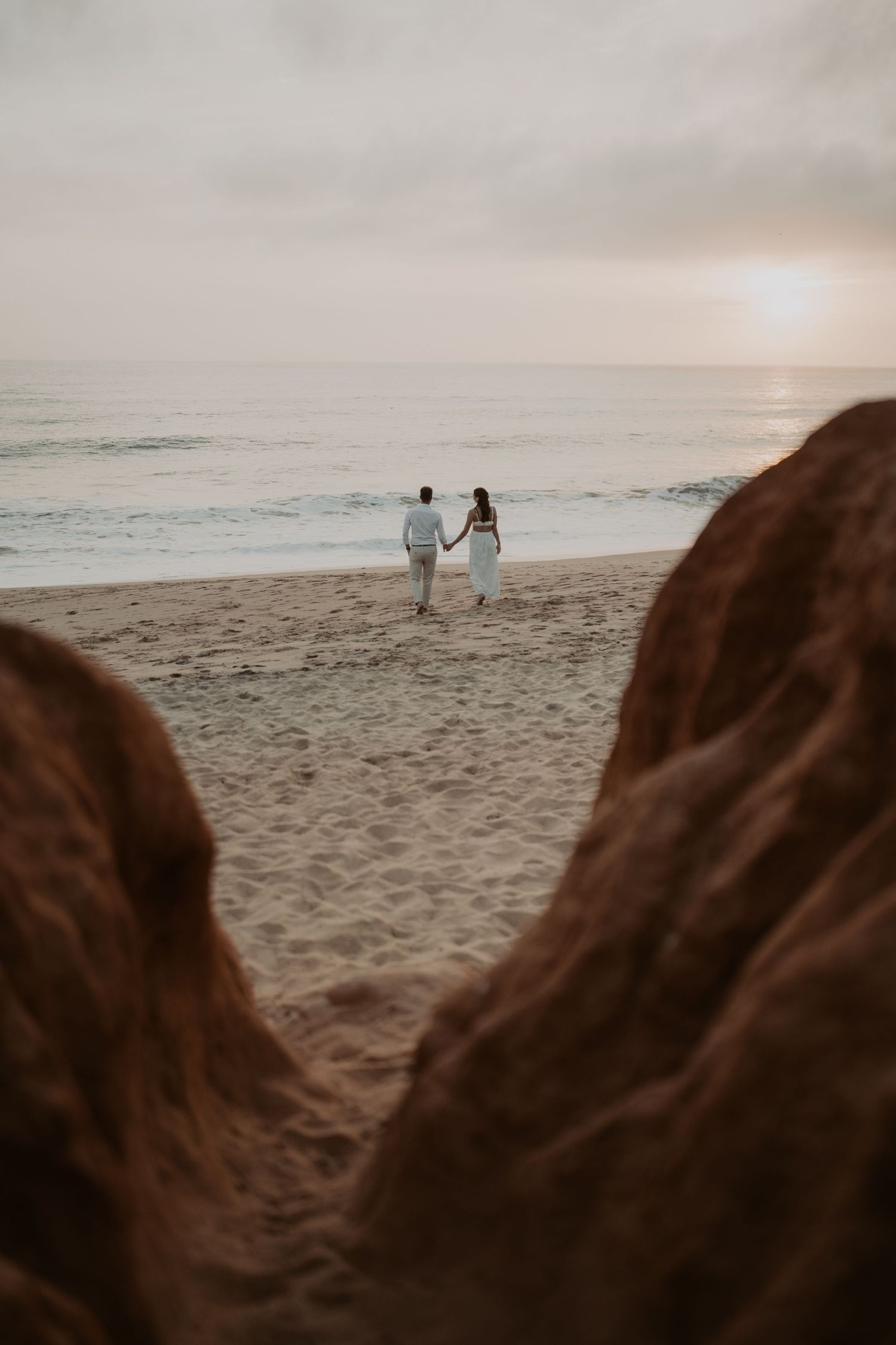 Couple in white walking hand-in-hand on a sandy beach at sunset, framed by rocky formations in the foreground.