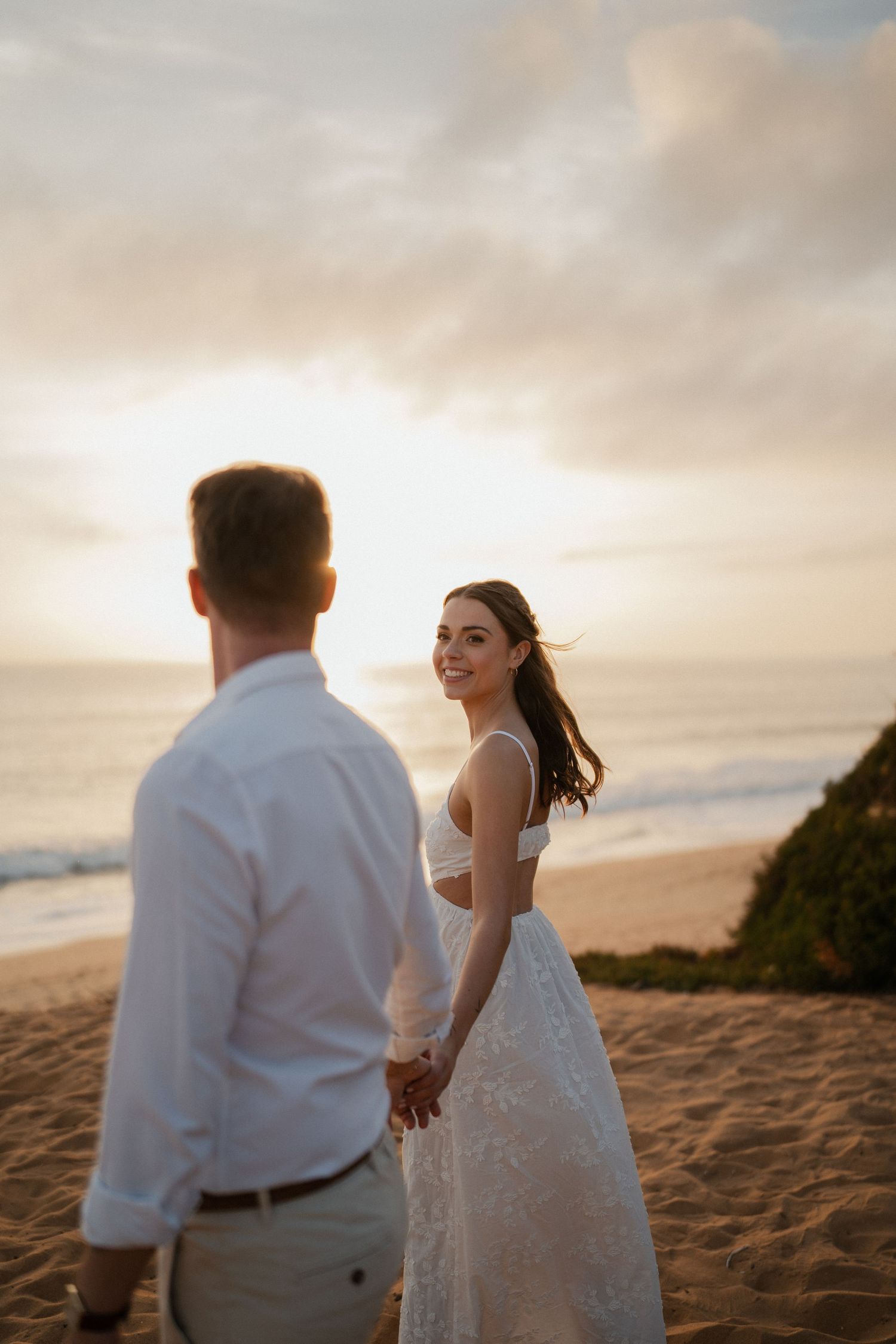 Bride in white lace dress smiles at groom on sandy beach during golden sunset wedding photoshoot.