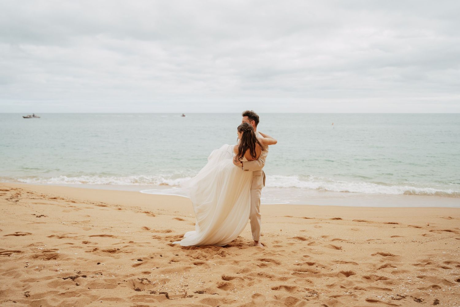 Couple in wedding attire embracing on a sandy beach with calm ocean waves and overcast sky backdrop.