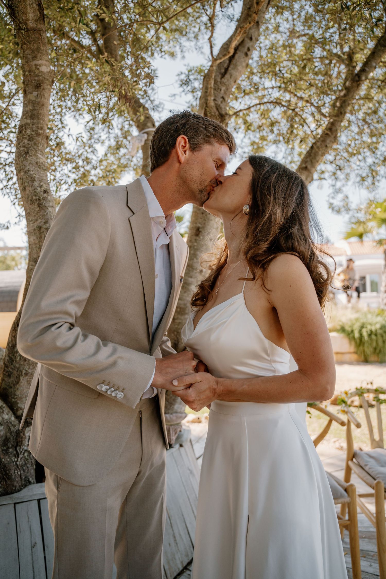 Bride and groom share first kiss under olive trees at outdoor wedding.