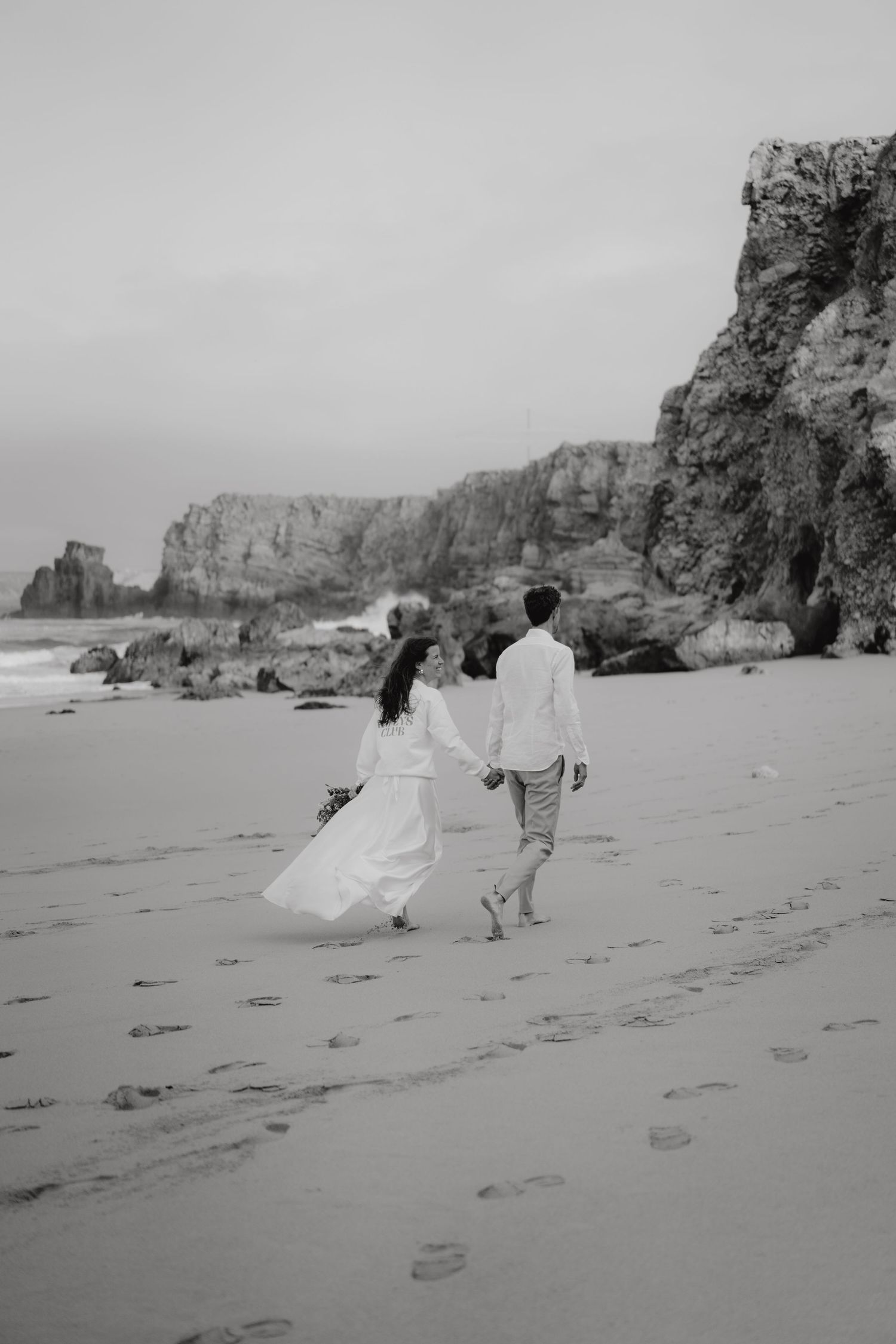 Black and white photo of bride and groom holding hands walking along a rocky beach shoreline.