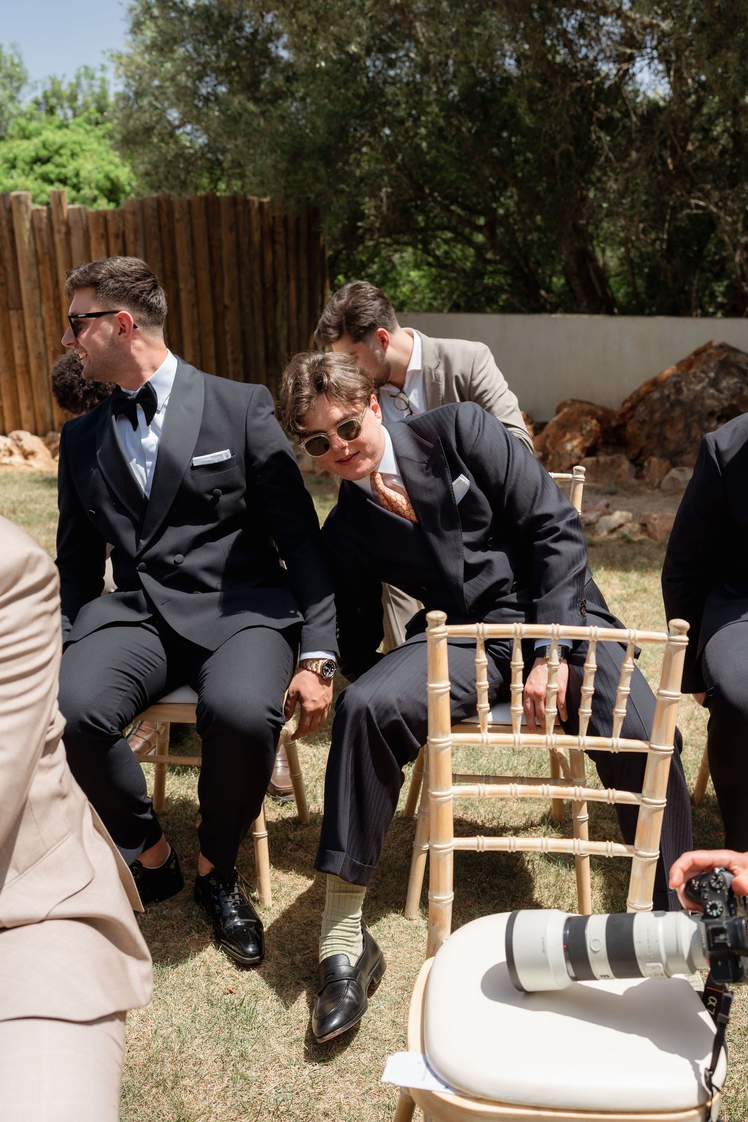 Men in suits sitting on wooden chairs at an outdoor wedding ceremony, one leaning back casually wearing sunglasses.