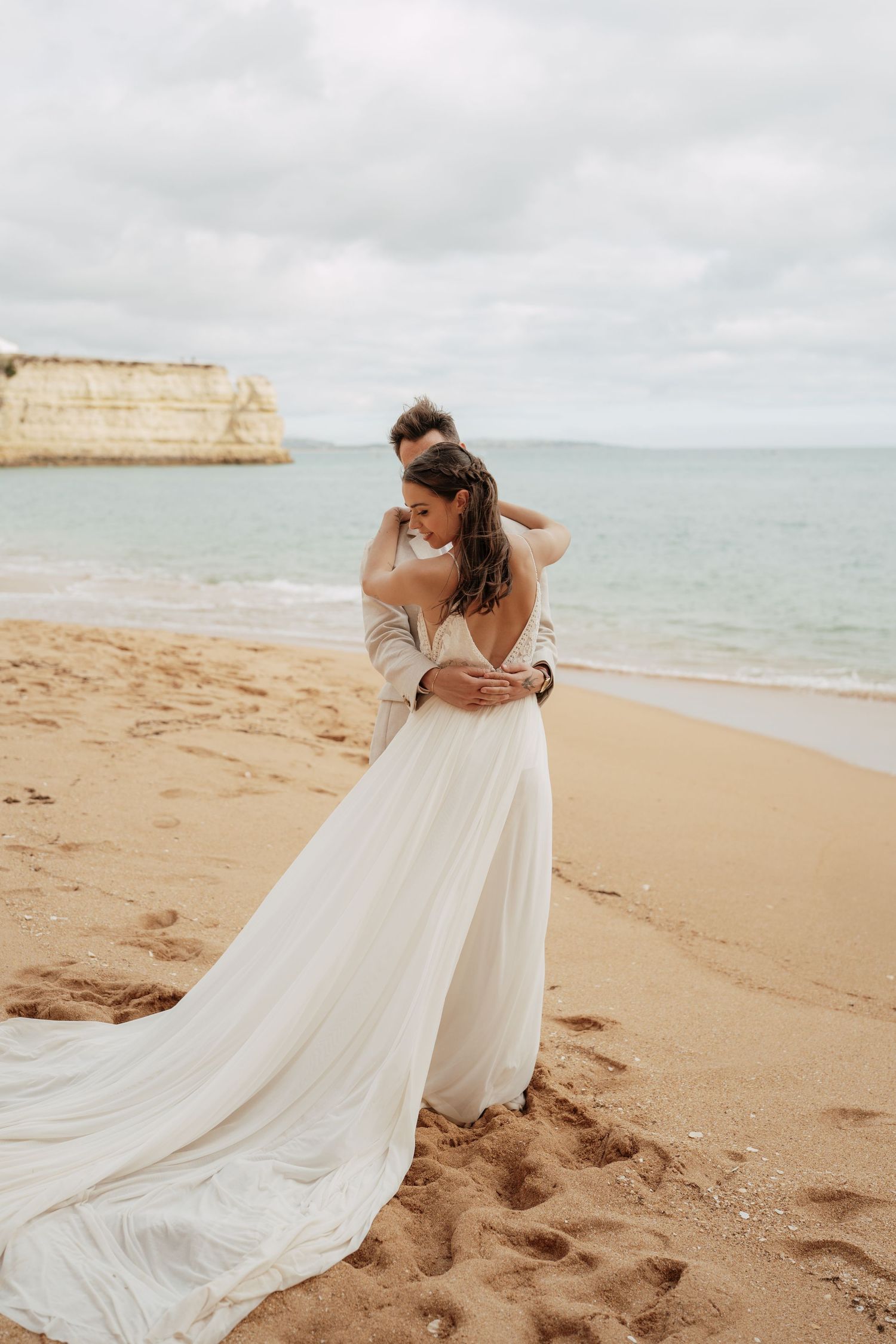 Bride and groom embracing on sandy beach in white wedding dress with long train, ocean and cliffs in background.