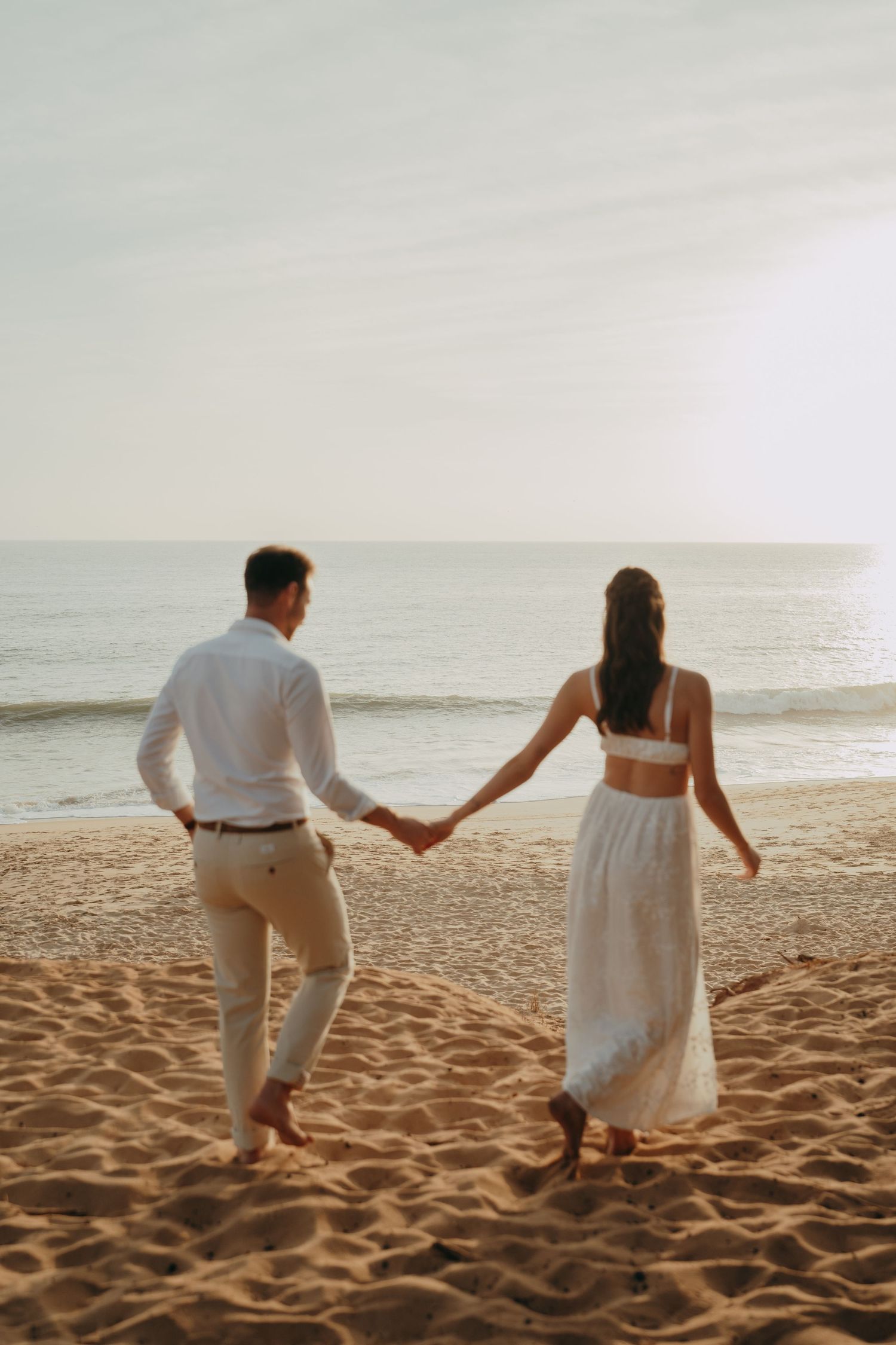 Couple holding hands walking on sandy beach at sunset, wearing white outfits, with ocean waves in the background.