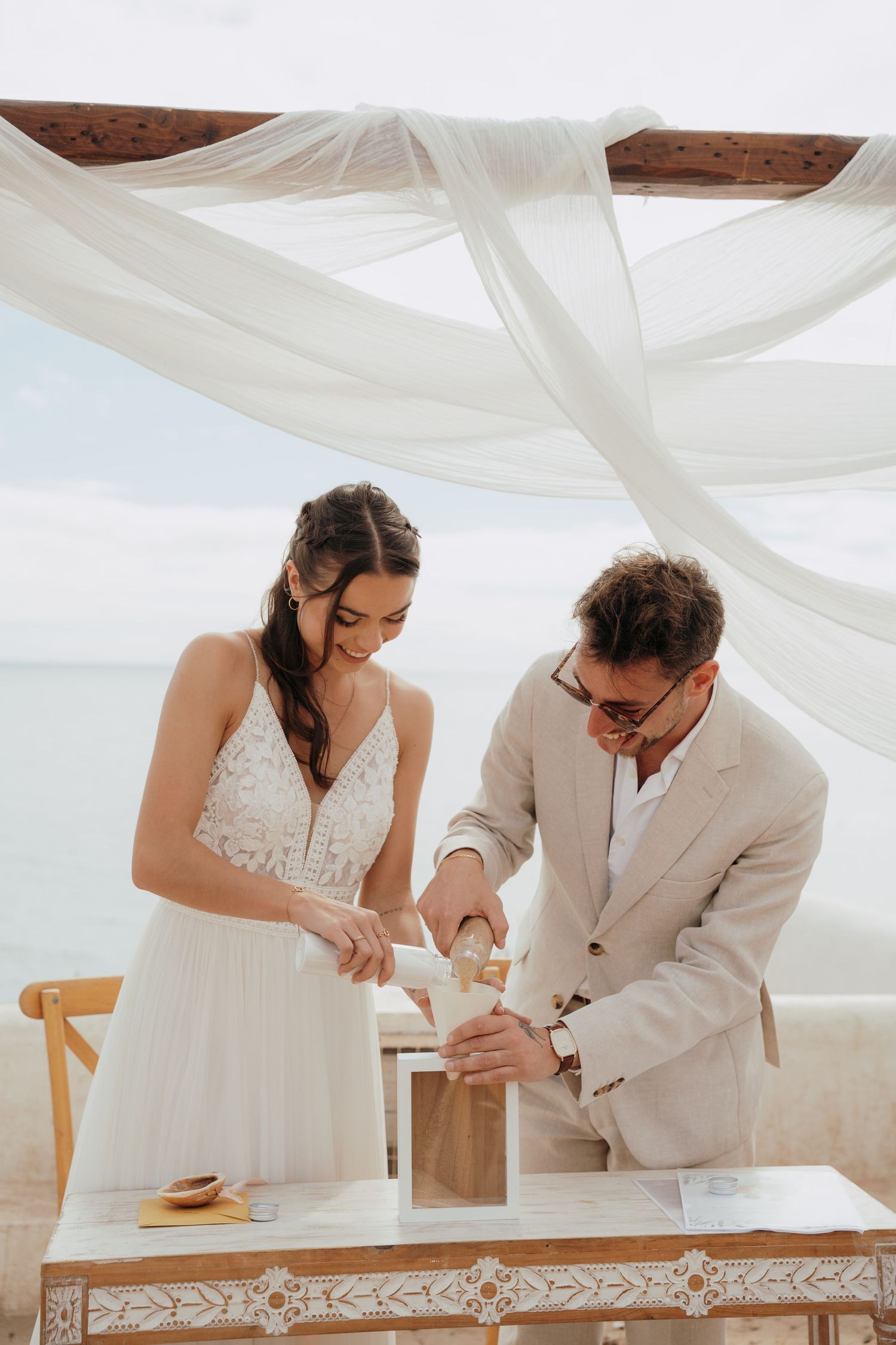 Bride and groom performing a sand ceremony at a beach wedding under a white draped canopy.