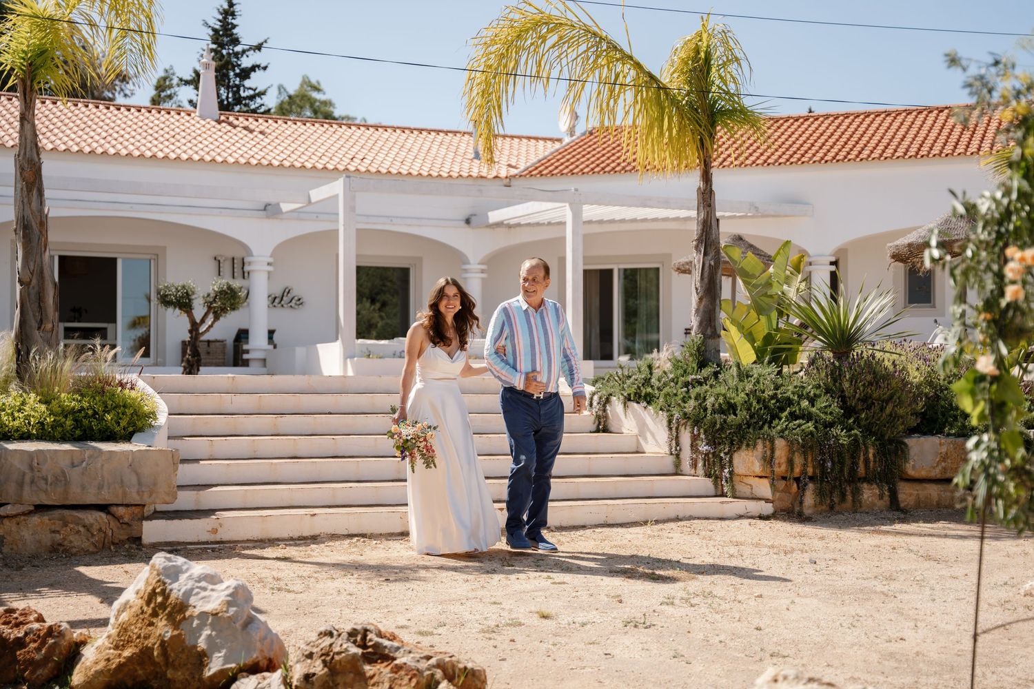 Bride and groom strolling in front of a white villa with palm trees.