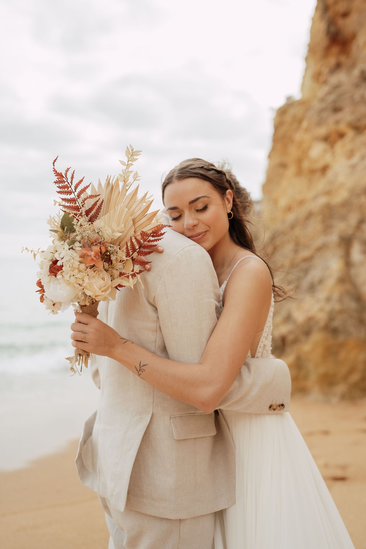 Bride and groom embrace on a sandy beach, she holds a boho bouquet with dried florals, rocky cliffs in the background.
