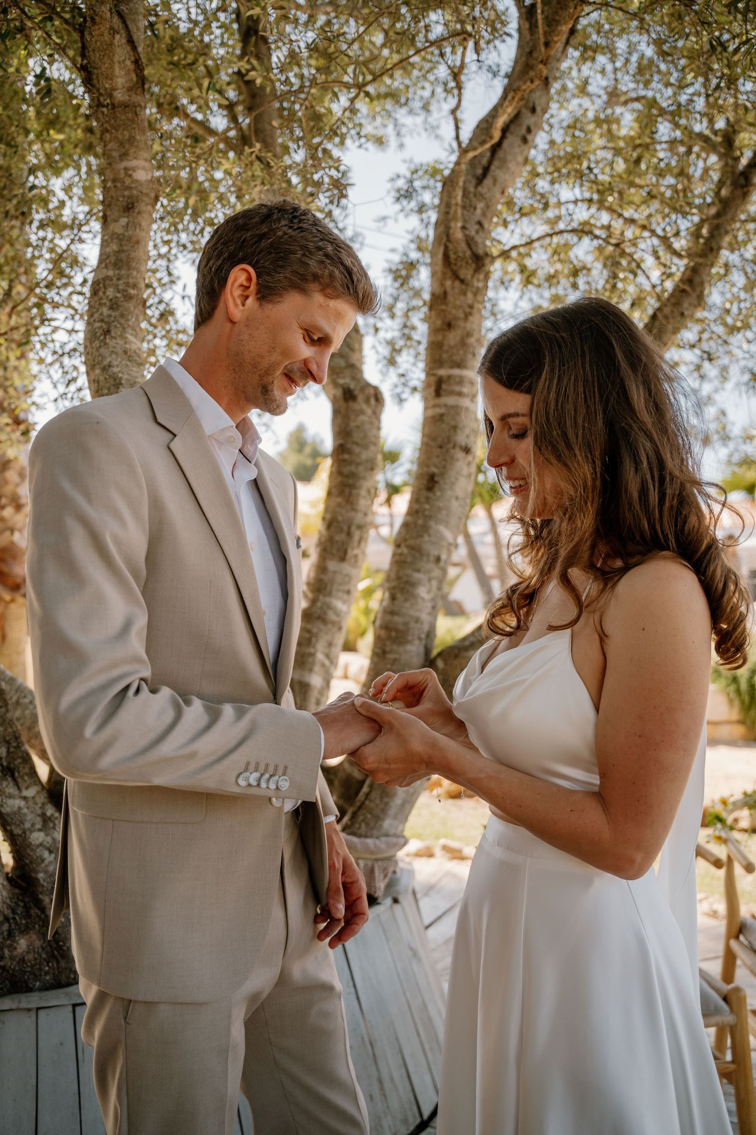 Groom places wedding ring on brides finger during sunny outdoor ceremony.