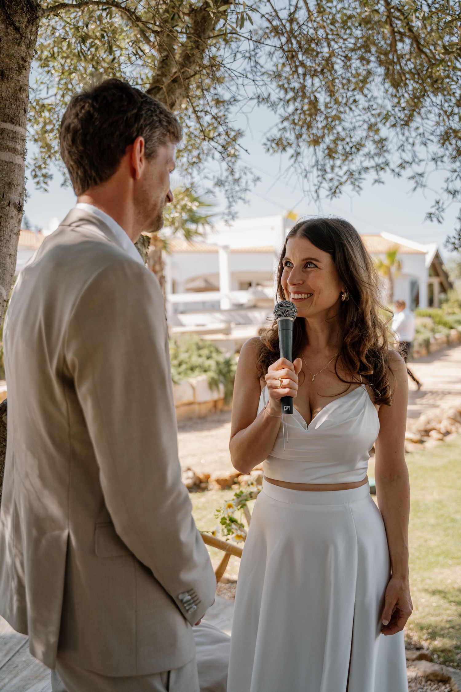 Smiling bride holds a microphone while reciting wedding vows to her groom under an olive tree.