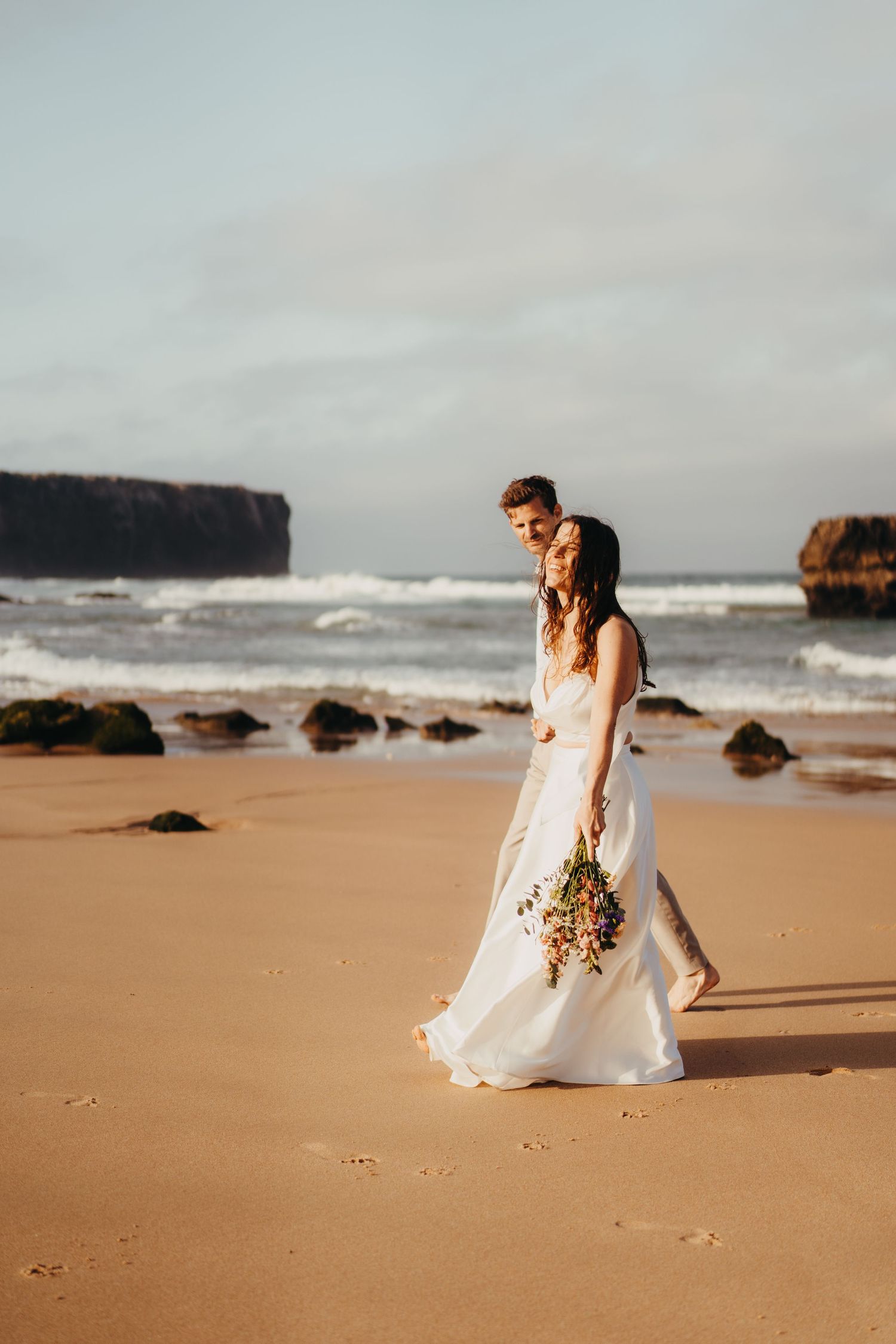 Groom holds bride from behind on a beach at sunset, she holds a colorful bouquet with ocean waves behind.