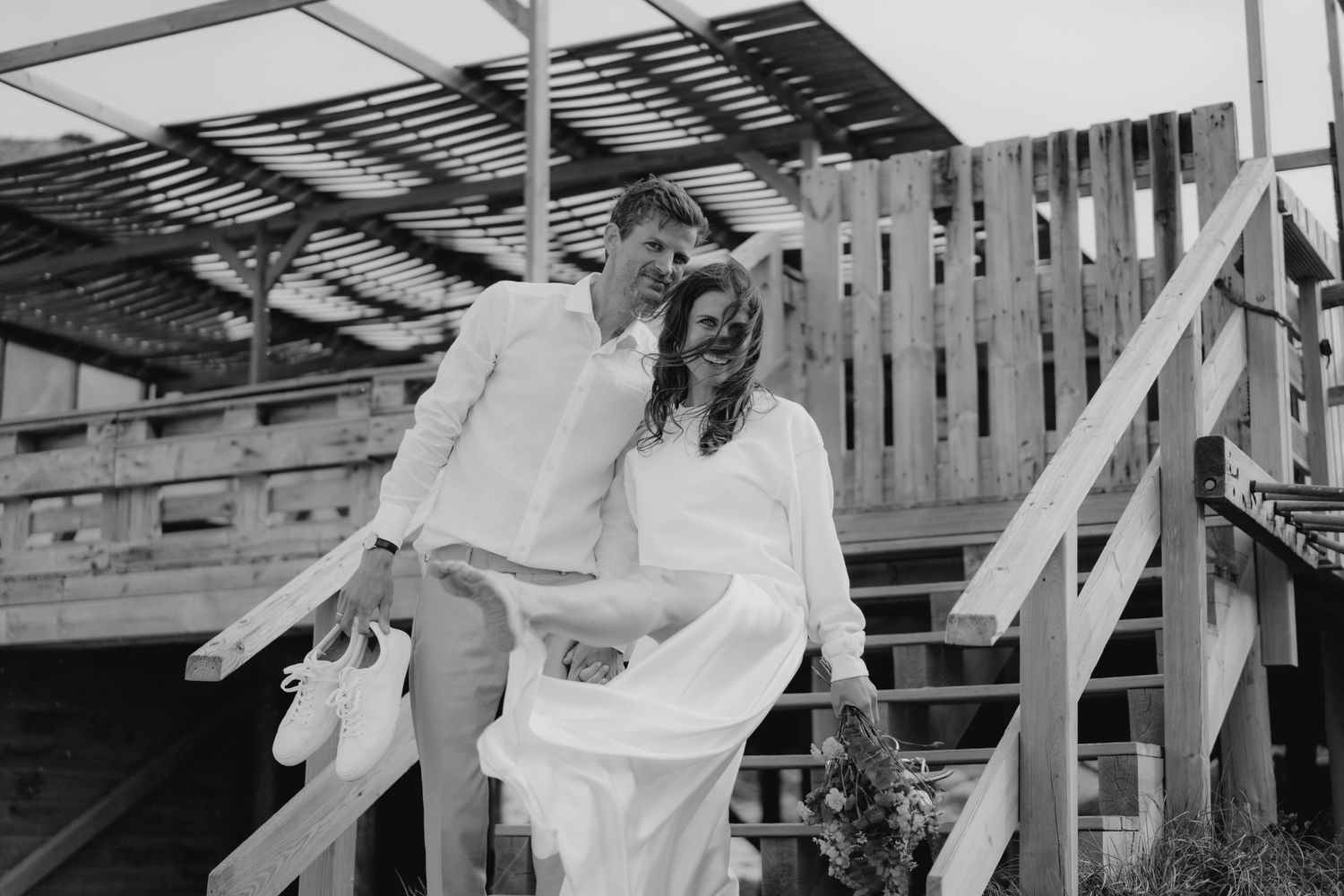 Black and white photo of a laughing bride in white dress and groom descending wooden stairs at their wedding.