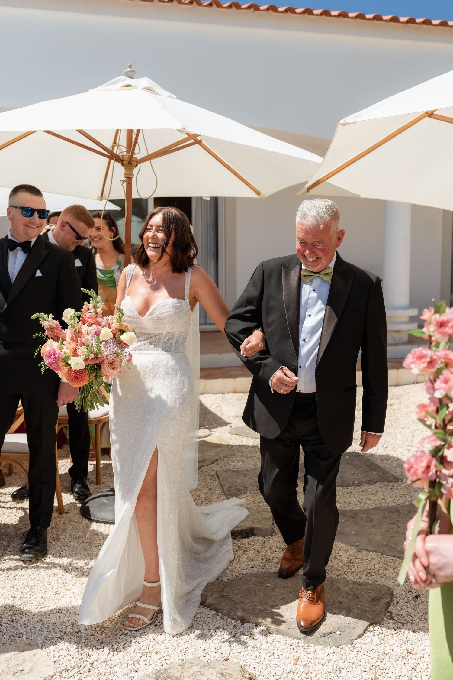 Bride in white gown walks with older gentleman down outdoor aisle, holding pink bouquet at sunny wedding.
