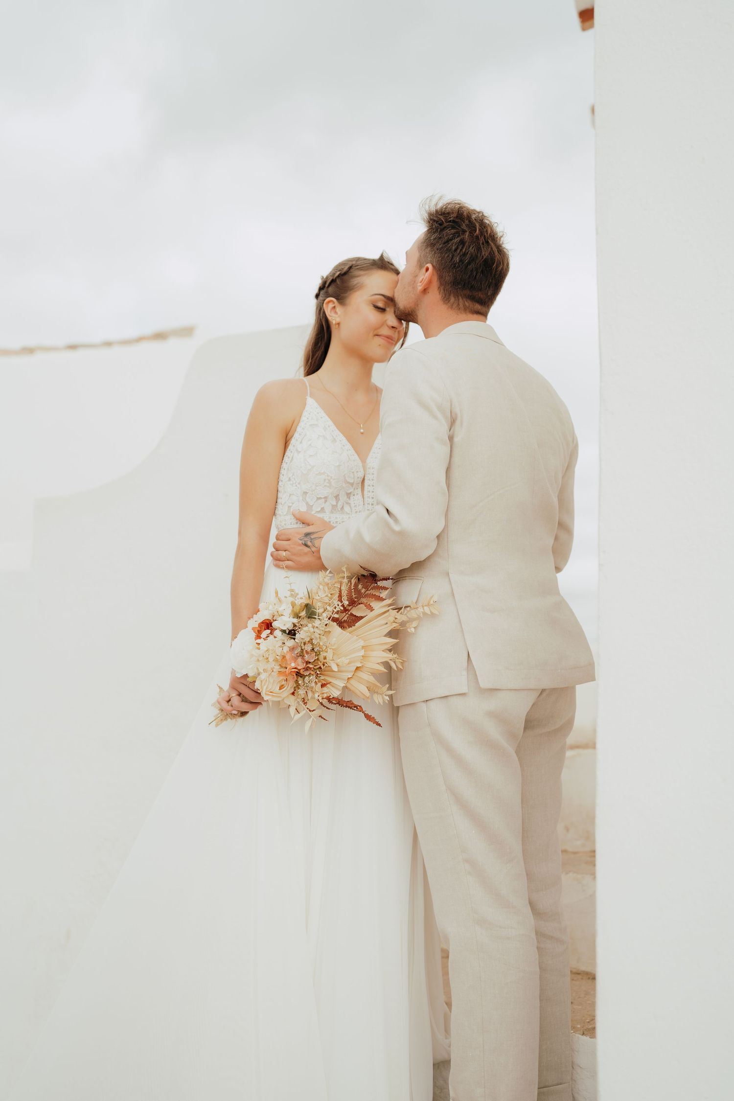 Bride and groom embrace tenderly outdoors, she holds a boho dried flower bouquet, both dressed in white and beige attire.