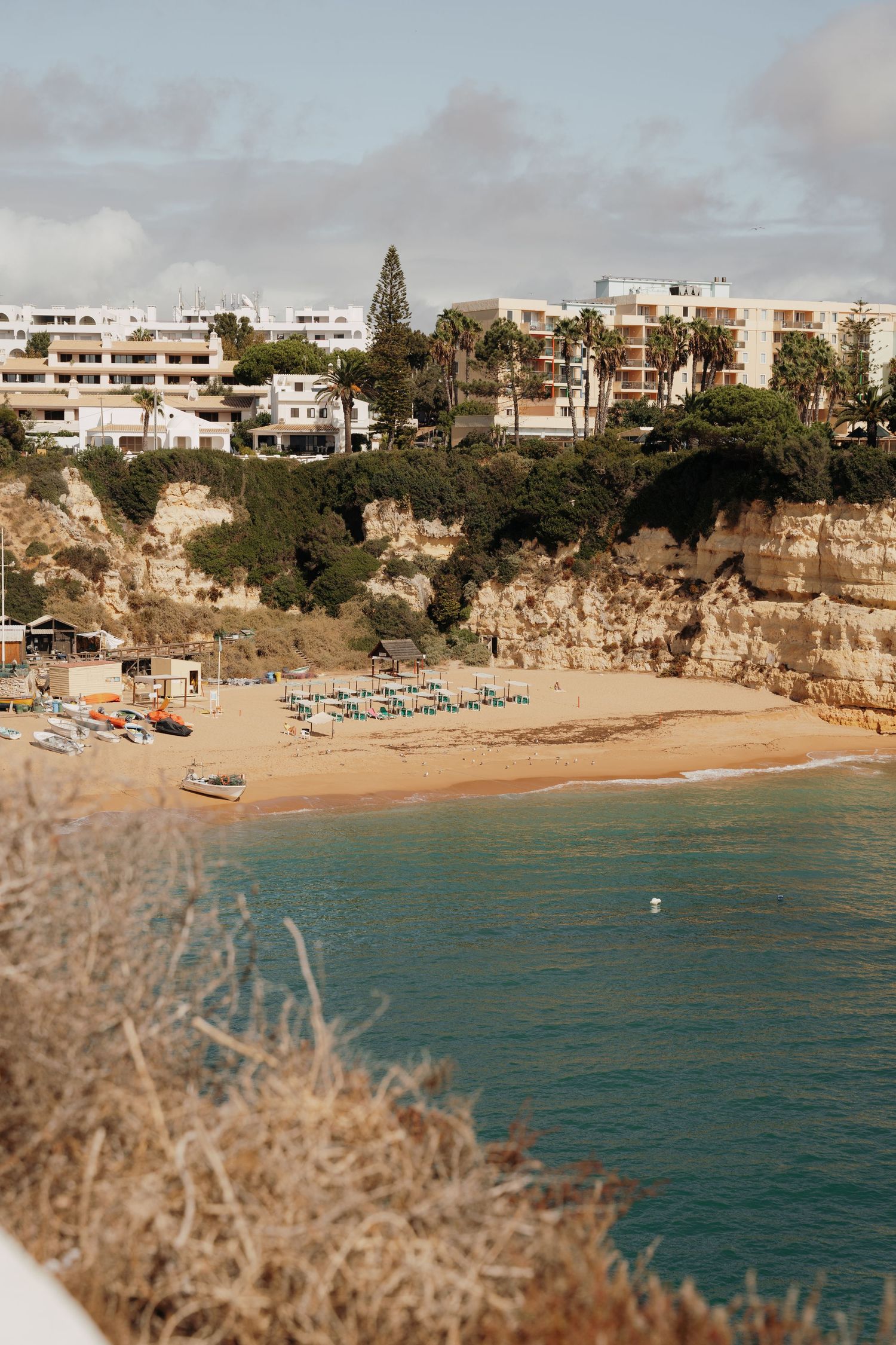 Sandy beach cove with turquoise water, golden cliffs, beach chairs, and white resort buildings surrounded by palm trees.