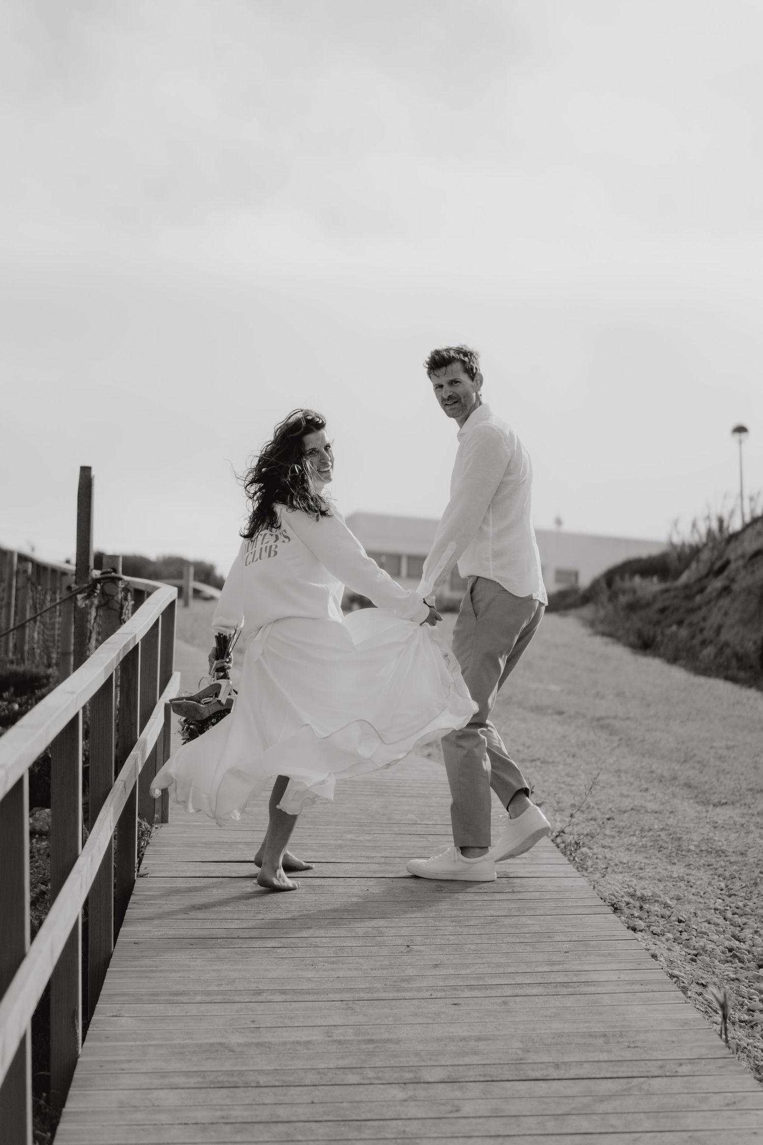 Black and white photo of bride and groom dancing on a wooden boardwalk near a sandy beach.