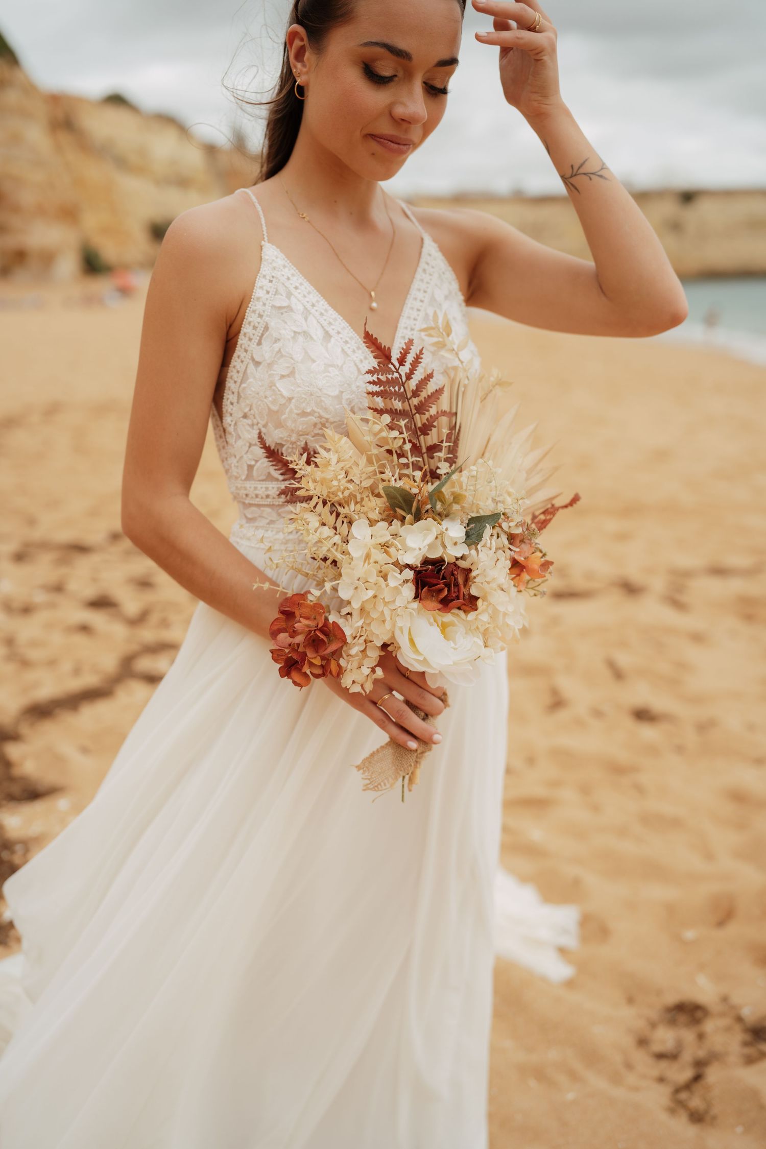 Bride in white lace gown holding boho bouquet with dried ferns and rust flowers on sandy beach.