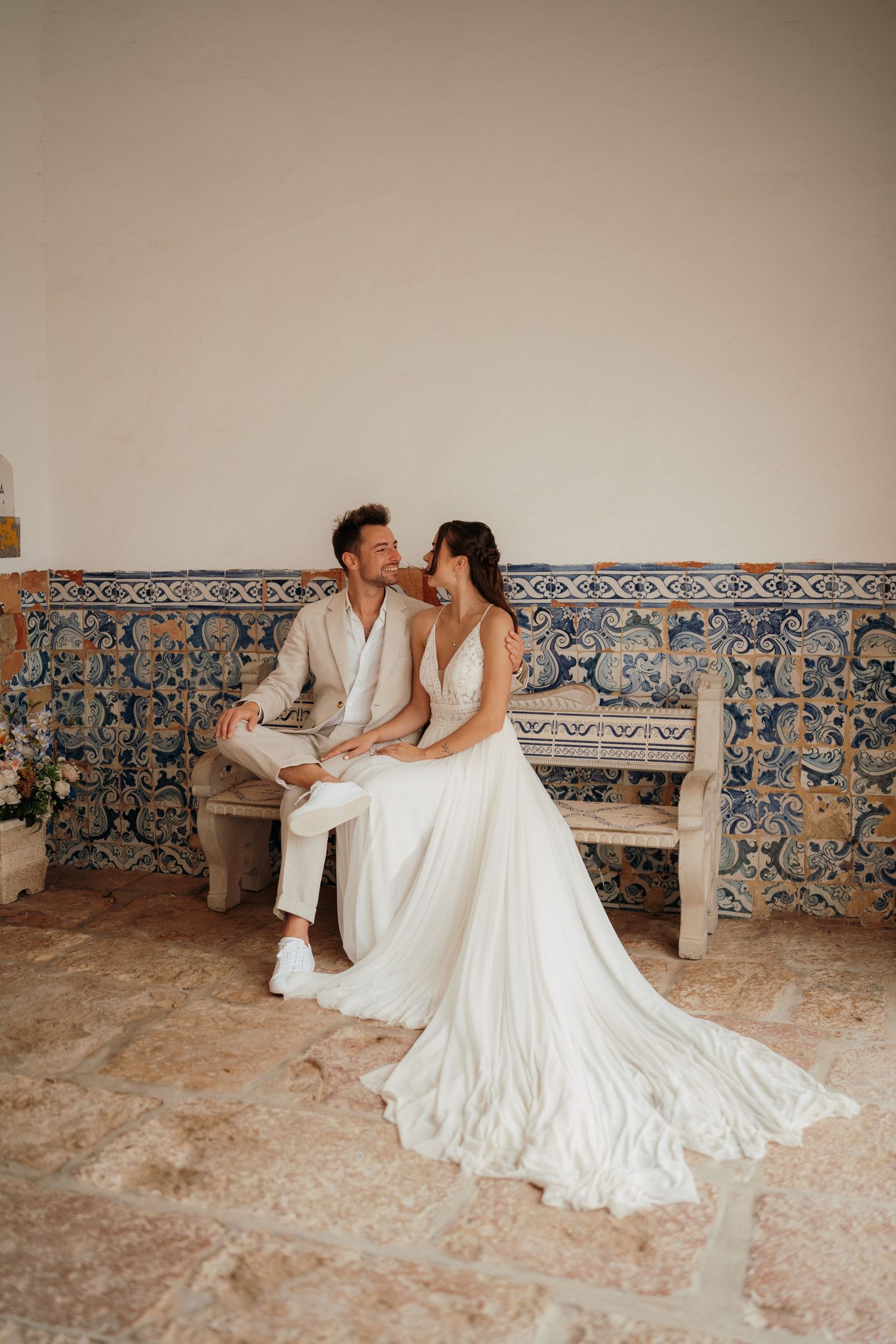 Bride and groom sitting together on a bench with blue azulejo tiles, sharing a romantic moment in their wedding attire.