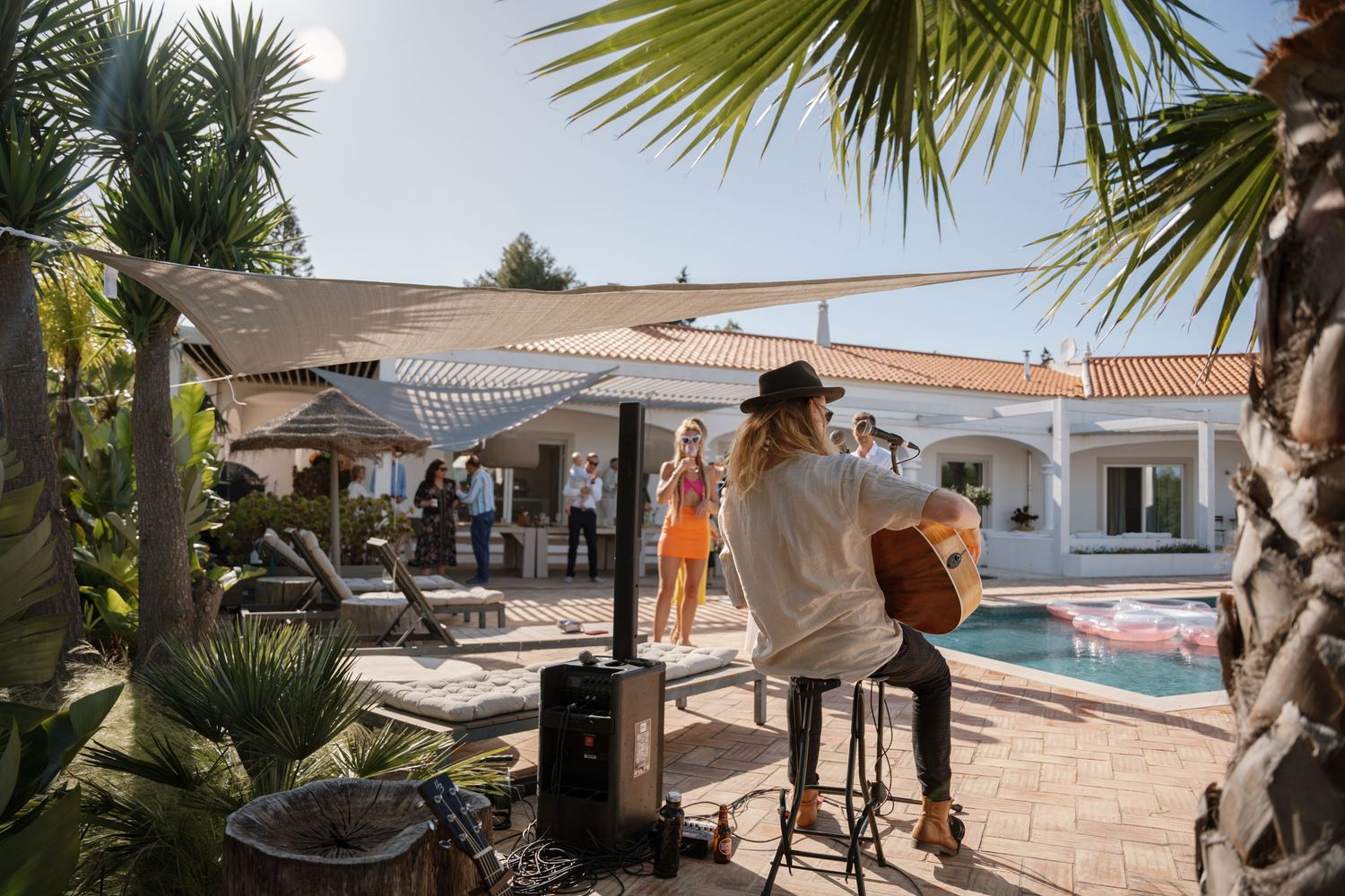 Acoustic guitarist performs poolside at an outdoor wedding reception surrounded by palm trees.
