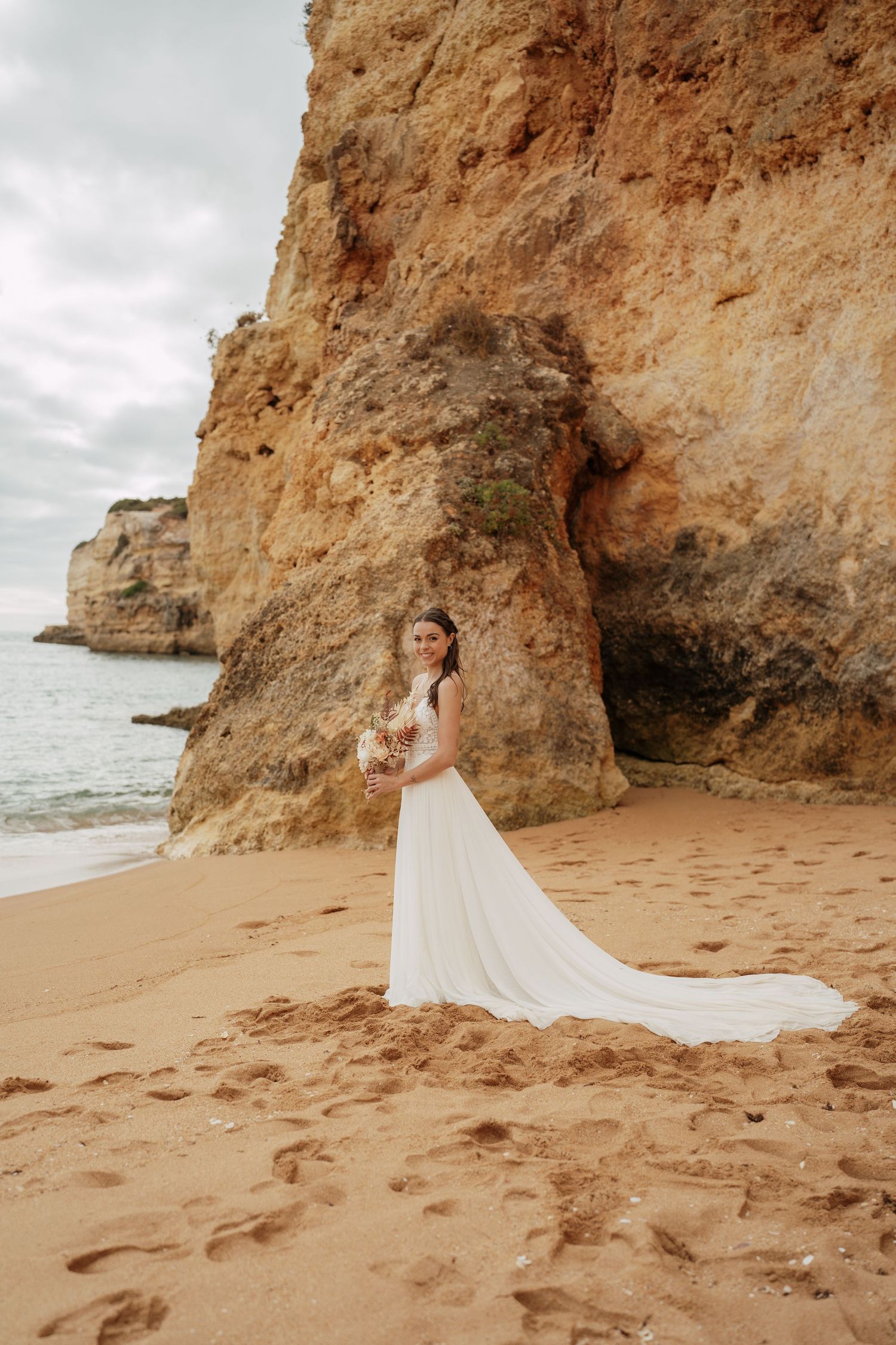 Bride in white gown with train stands on sandy beach beside dramatic rocky cliffs with ocean backdrop.