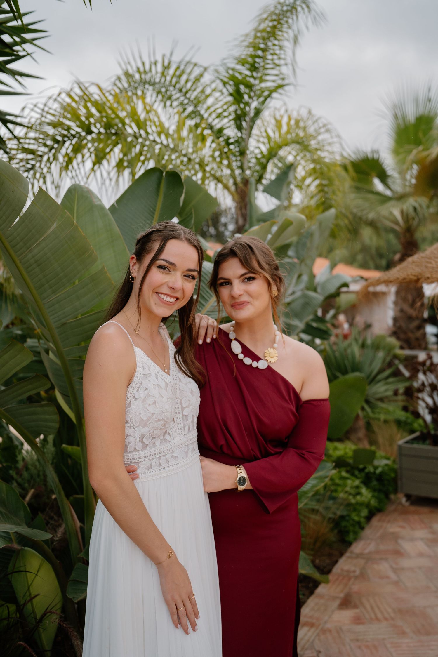 A bride in a white lace gown poses with a woman in a burgundy dress among tropical palms.
